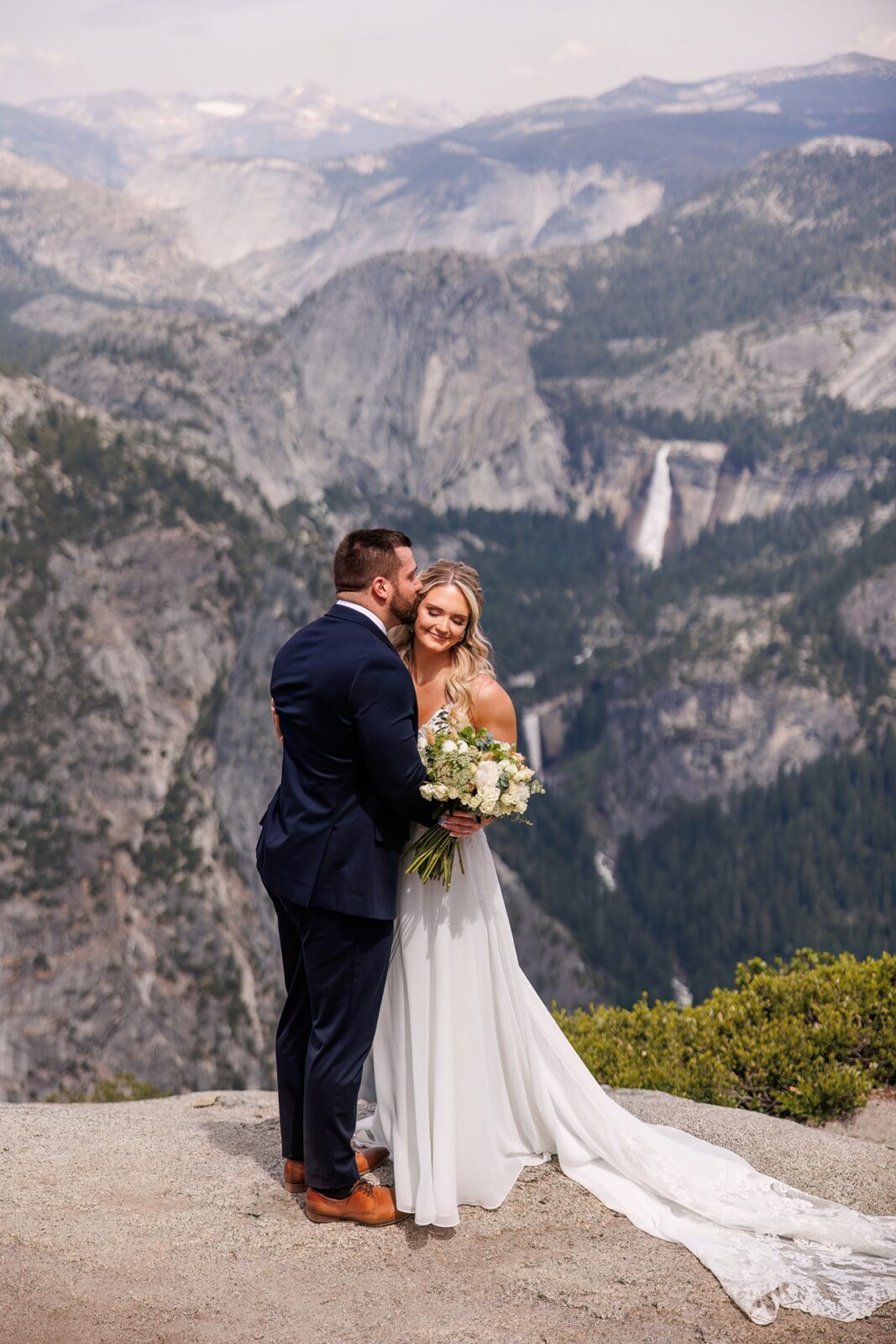 bride and groom cuddling at glacier point with yosemite waterfalls in the background 