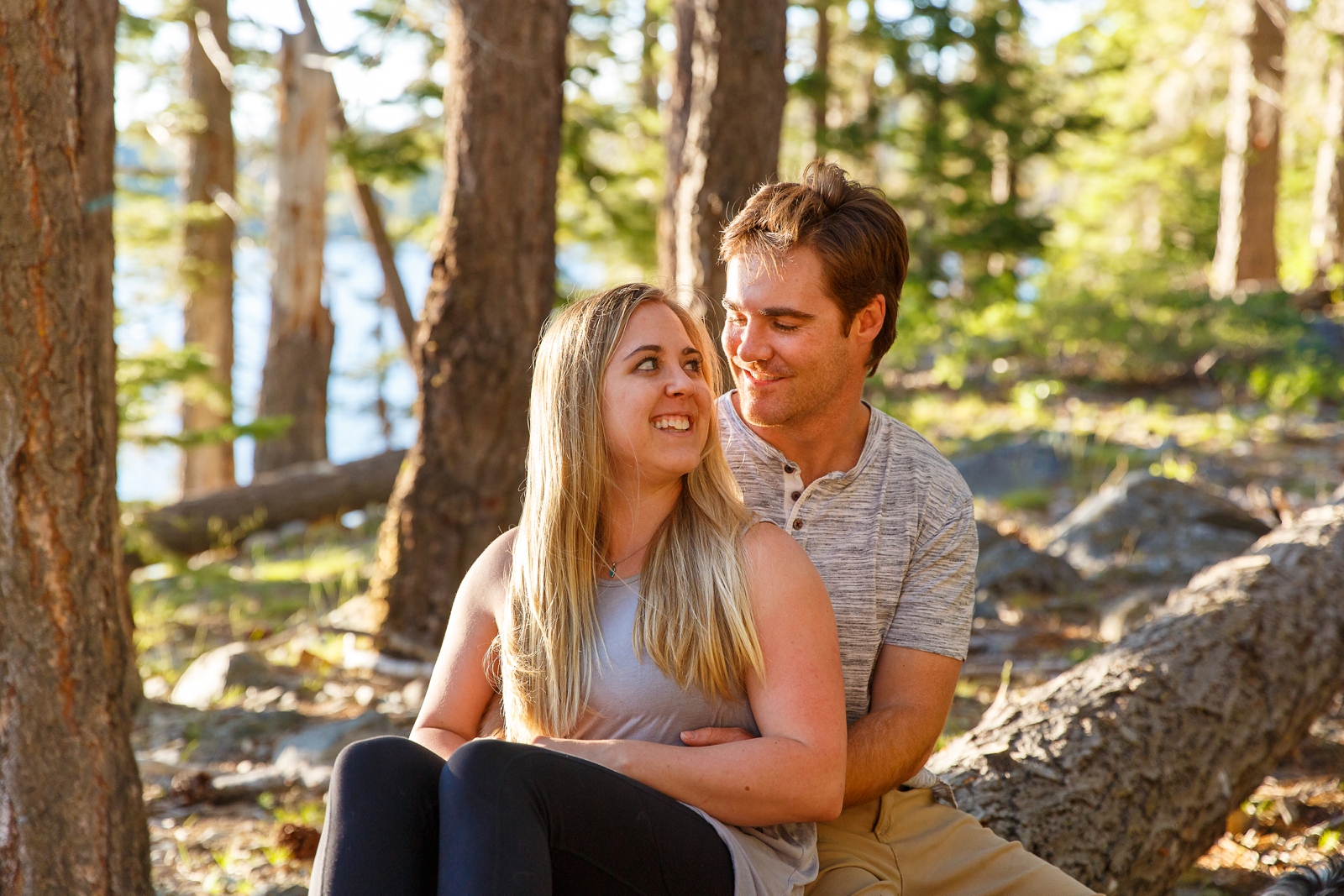 a couple sitting on a fallen branch with Lake Tahoe in the background looking at each other and smiling during their Tahoe area adventure session