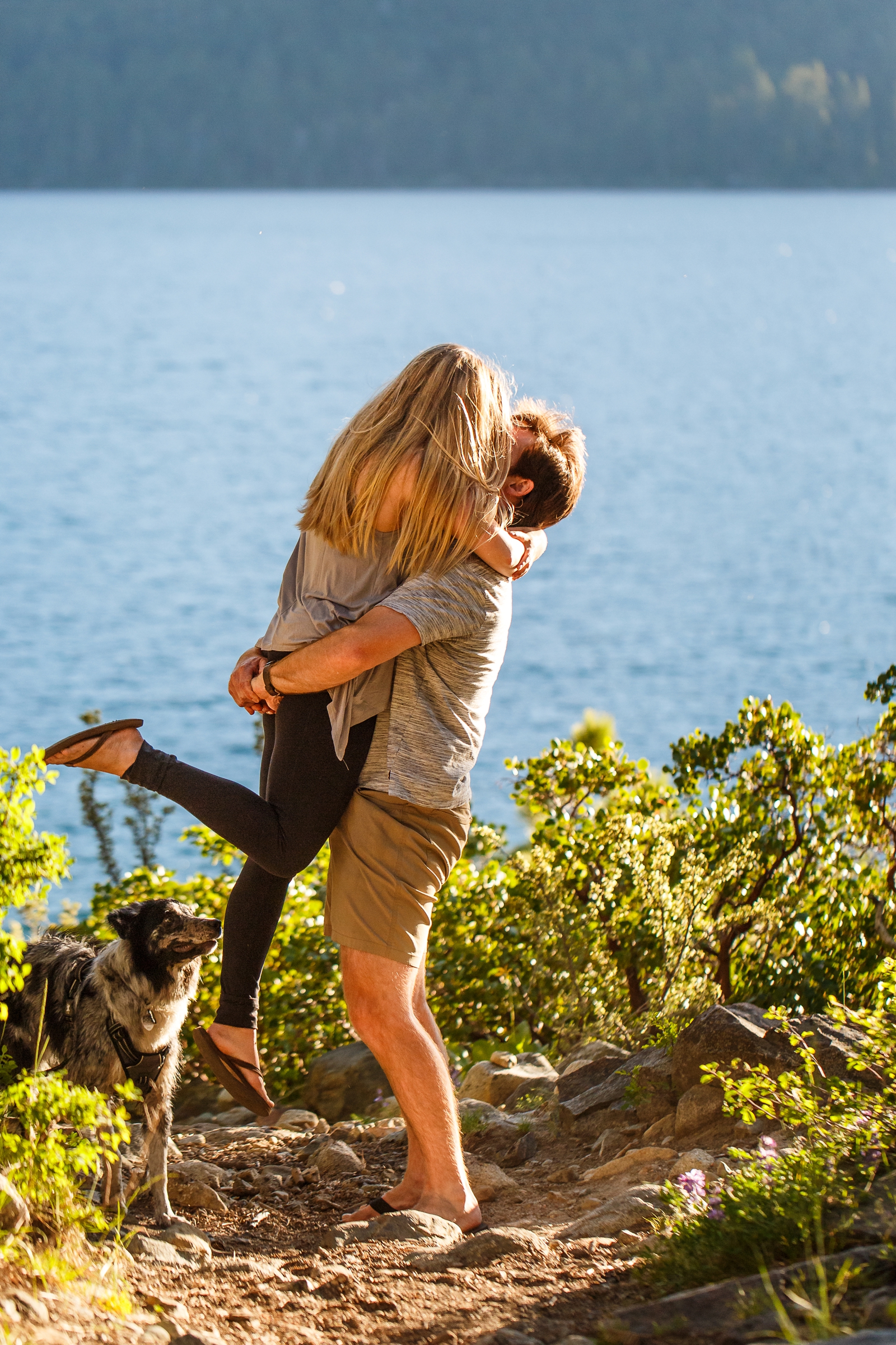 a man lifting his girlfriend and kissing her with their dog behind them by Lake Tahoe for their adventure session