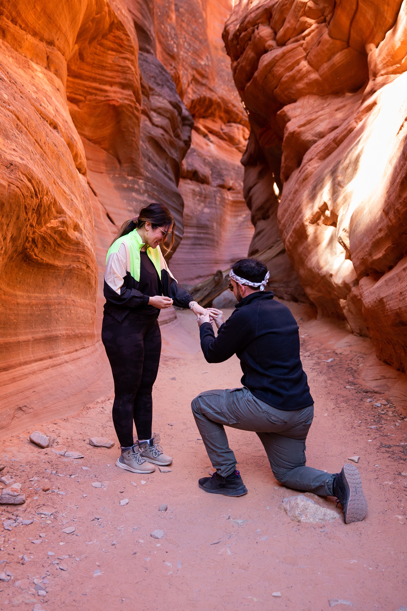 A guy down on his knee proposing to and putting the ring on his shocked and surprised girlfriend in the middle of the Utah slot canyon during their adventurous hike