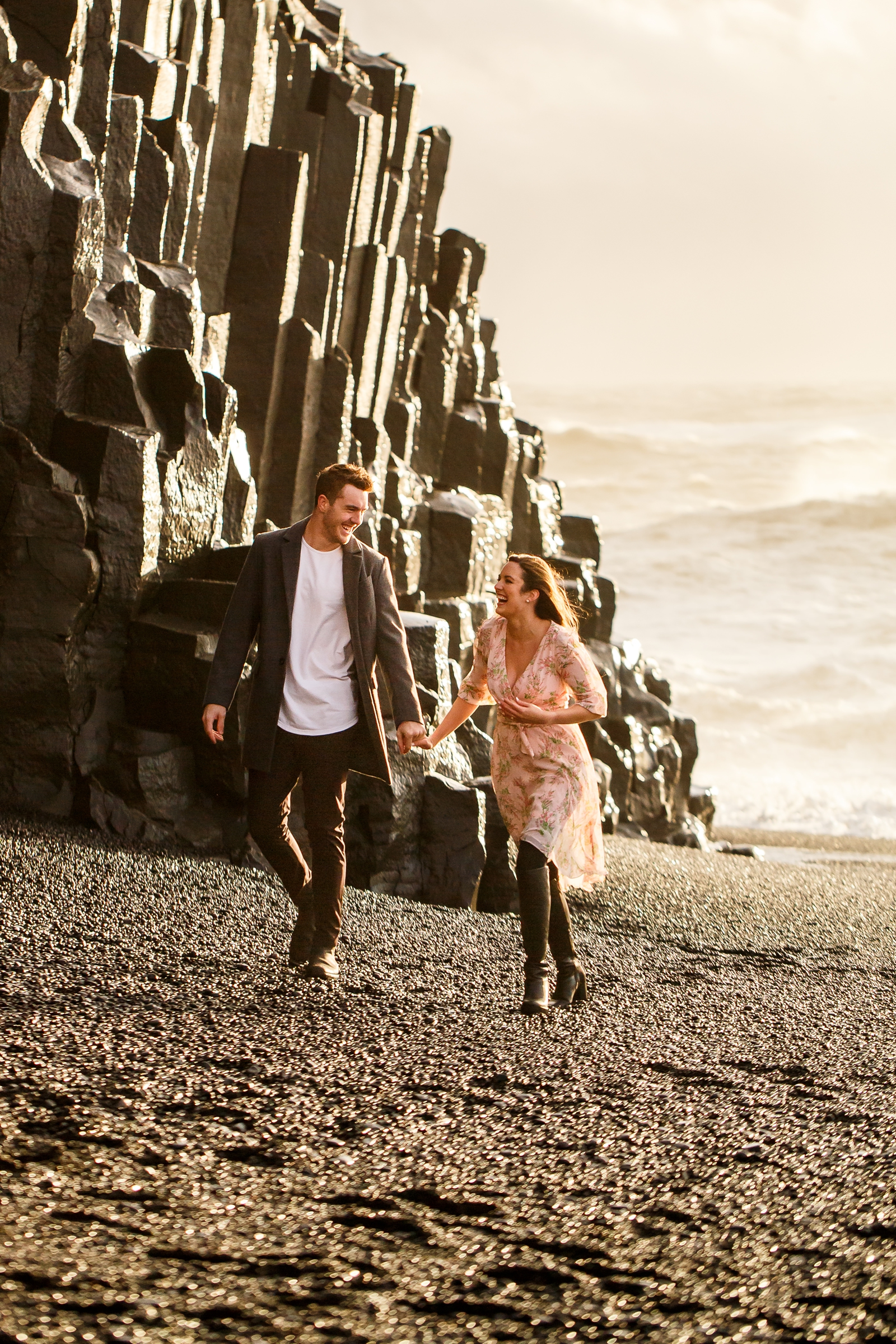 Engaged Couple looking at each other and holding hands walking on the black sand beach by the basalt columns at sunset with the ocean in the background at their destination Iceland engagement session near Vik, Iceland