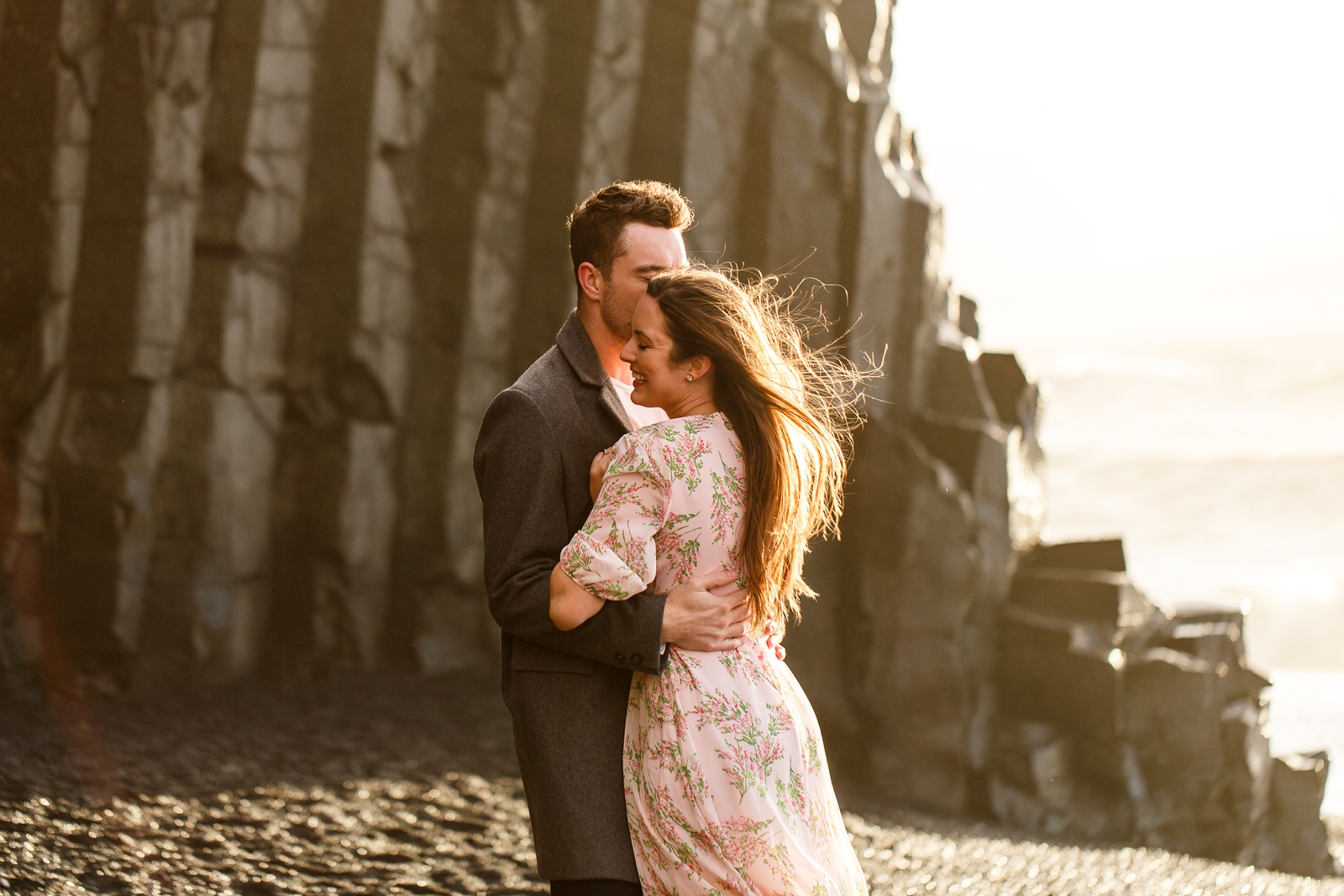 Engaged Couple looking at each other and snuggling by the basalt columns with the ocean in the background at their destination Iceland engagement session near Vik, Iceland