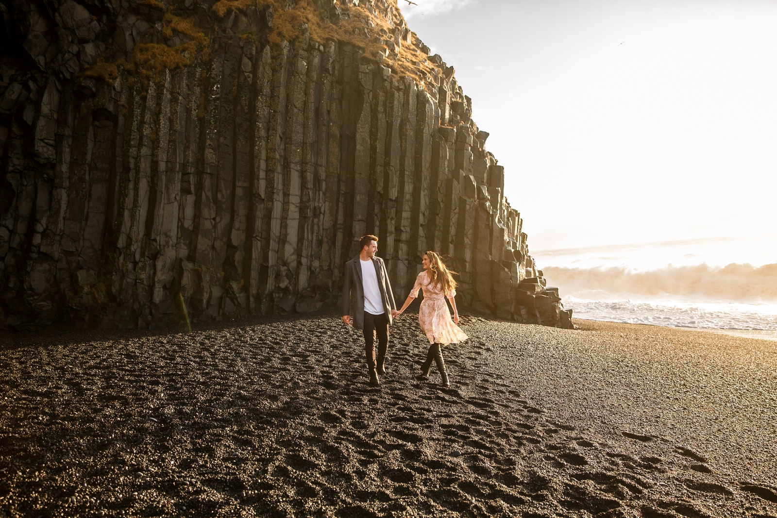 Engaged Couple holding hands walking on the black sand beach by the basalt columns with the ocean in the background at their destination Iceland engagement session near Vik, Iceland