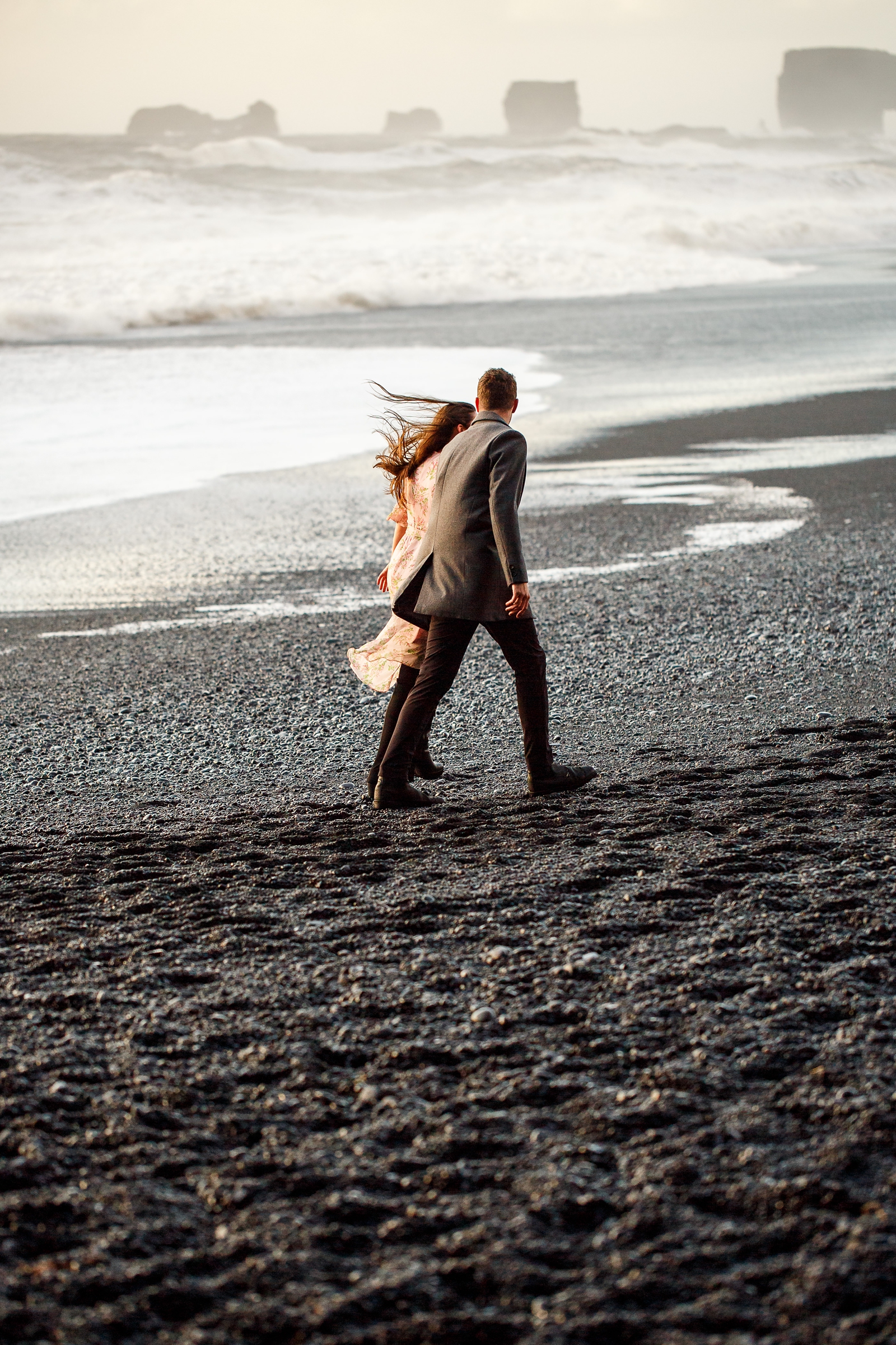 Engaged Couple holding hands walking on the black sand beach by the basalt columns with the ocean in the background at their destination Iceland engagement session near Vik, Iceland