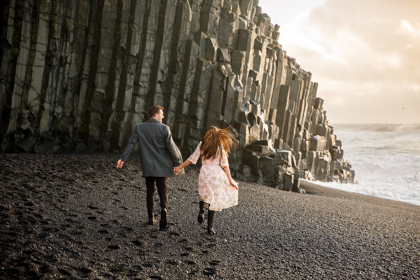 Engaged Couple holding hands running on the black sand beach by the basalt columns with the ocean in the background at their destination Iceland engagement session near Vik, Iceland