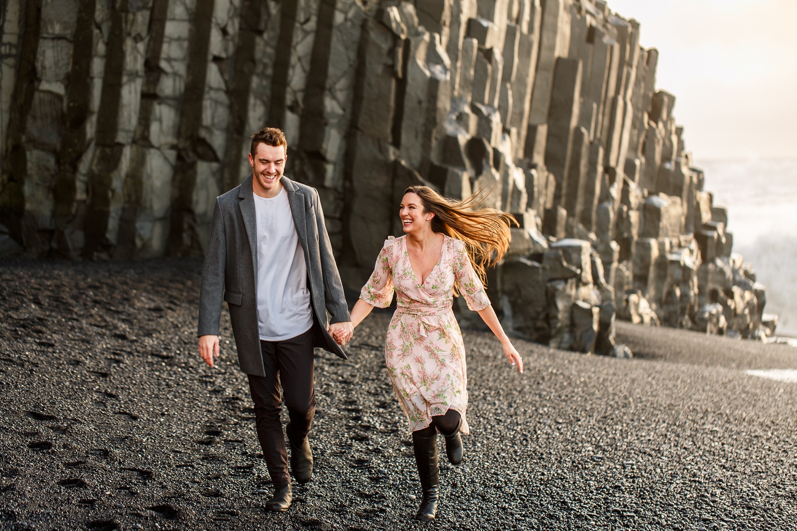 Engaged Couple holding hands running on the black sand beach by the basalt columns with the ocean in the background at their destination Iceland engagement session near Vik, Iceland