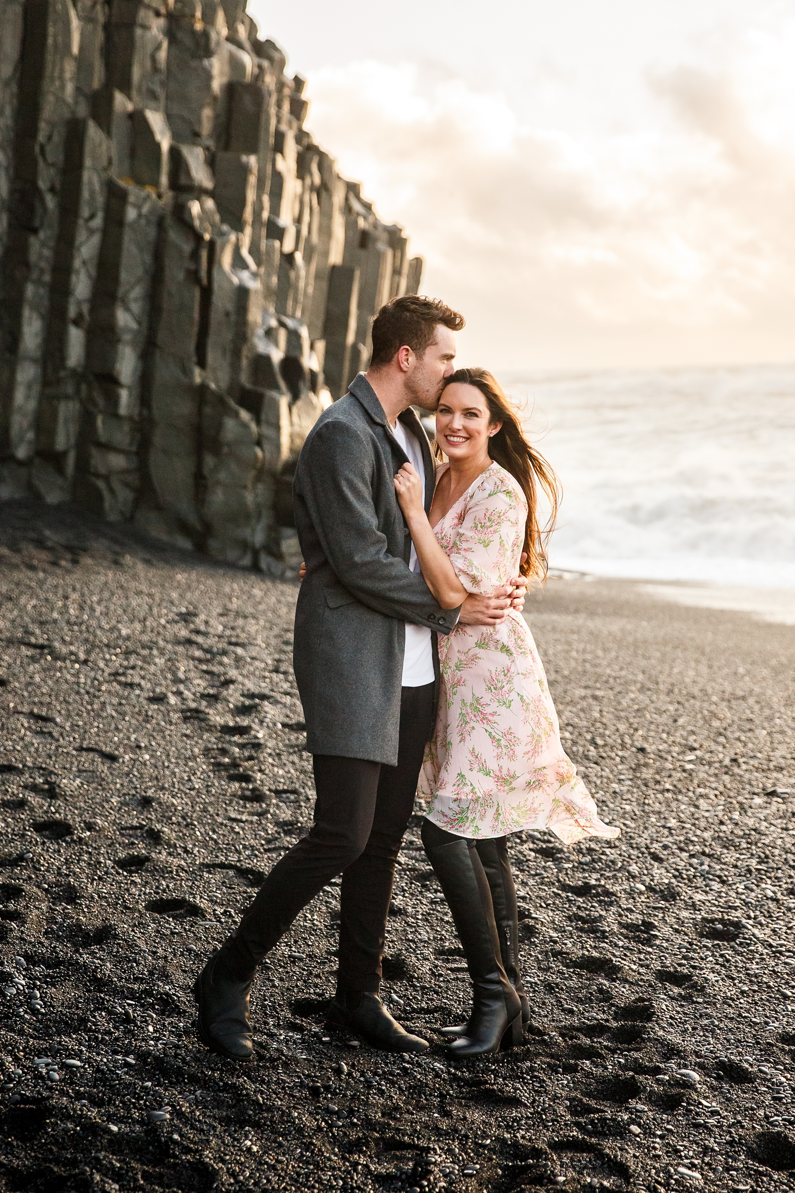 Engaged Couple smiling with her fiance kissing her on the forehead by the basalt columns with the ocean in the background at their destination Iceland engagement session near Vik, Iceland