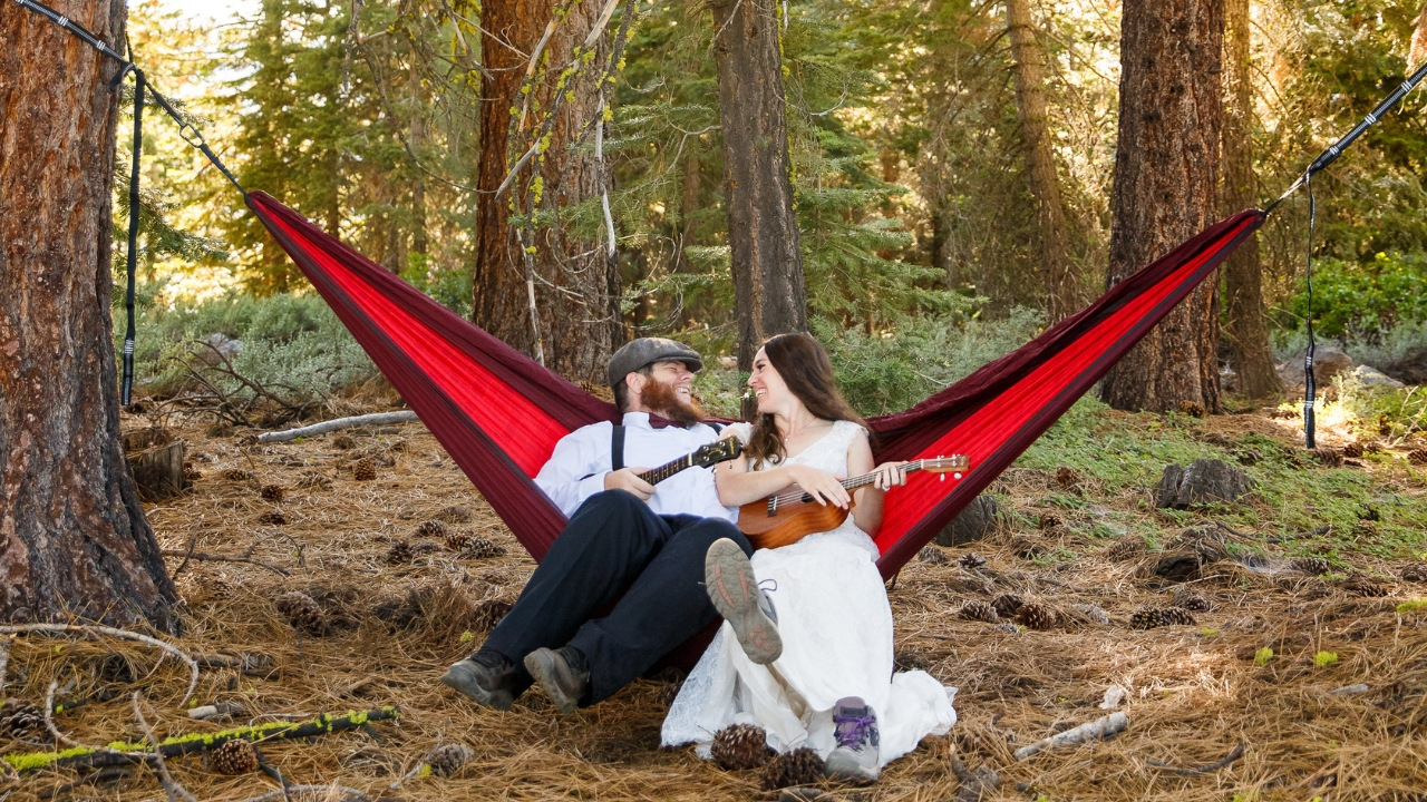 photo of bride and groom sitting on a hammock in the woods playing banjos