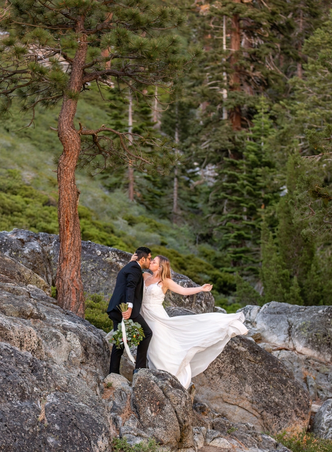 Photo of bride and groom kissing on rocky ledge near a tree as her dress blows in the wind