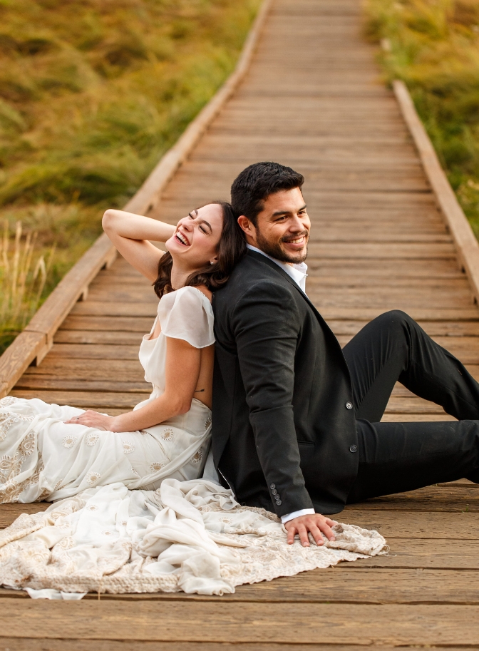 photo of bride and groom sitting on wooden pathway back to back laughing