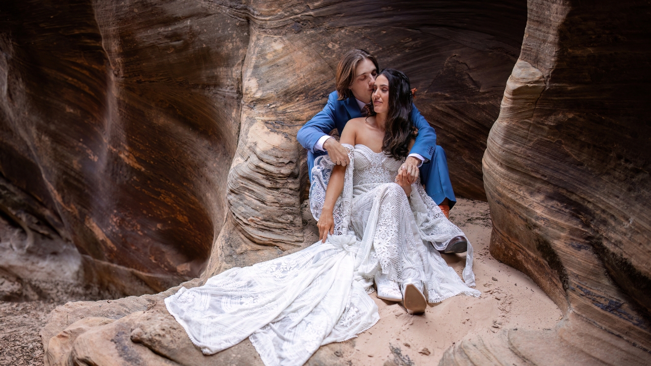 photo of groom kissing bride's cheek while sitting down in a slot canyon in Zion National Park