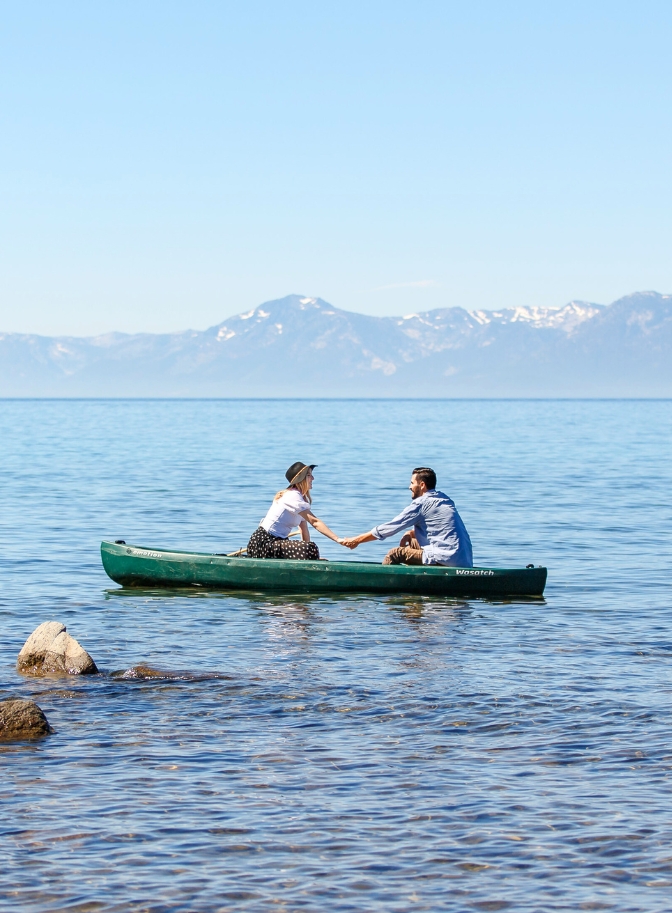 photo of couple holding hand in canoe with mountain range behind them