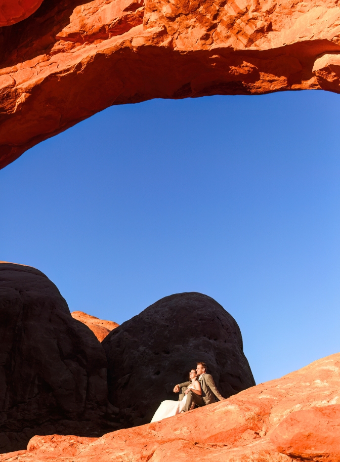 photo of bride and groom sitting down on rockface to watch sunset at Arches National Park