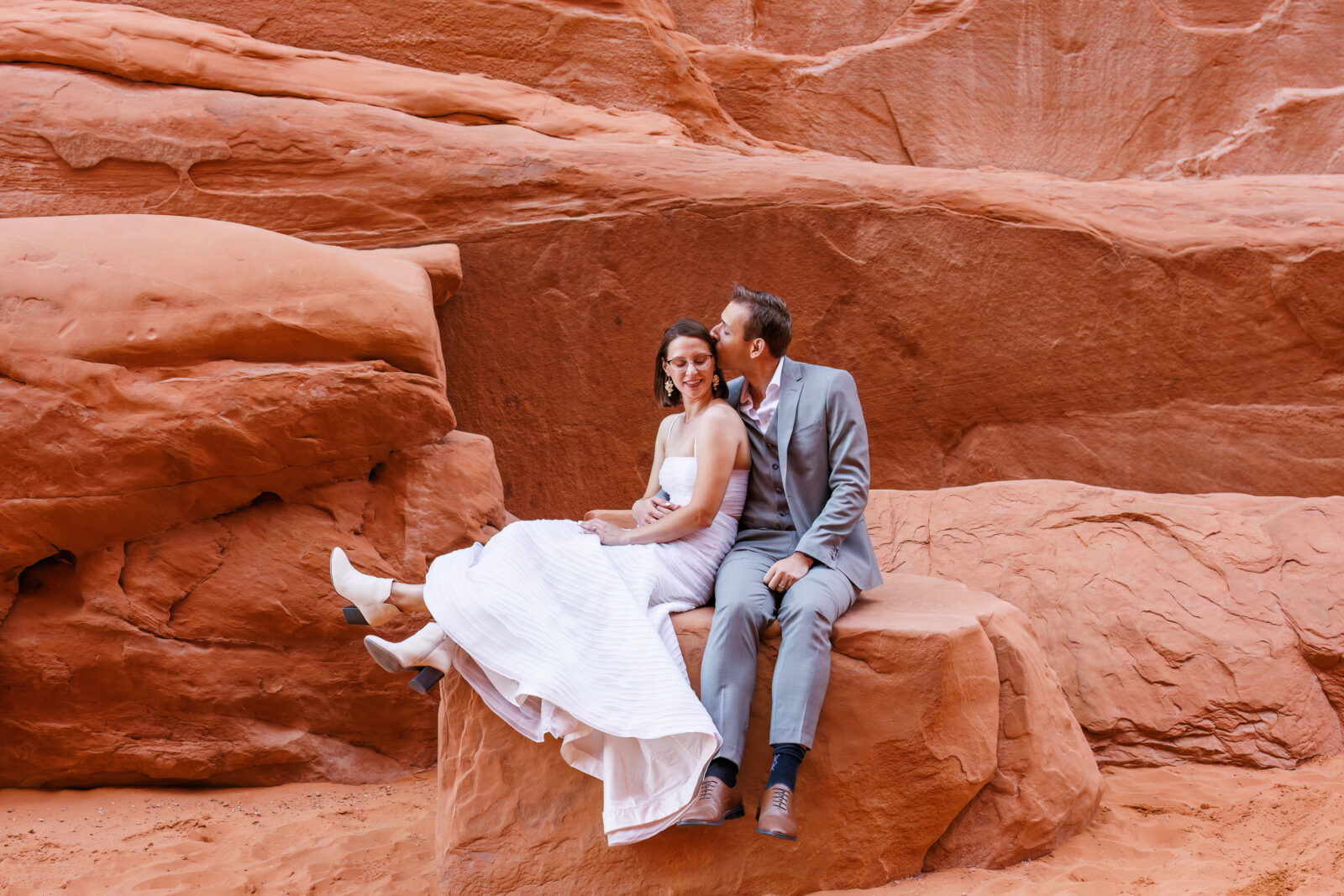 photo of bride and groom sitting on rock embracing while groom kisses bride on the forehead