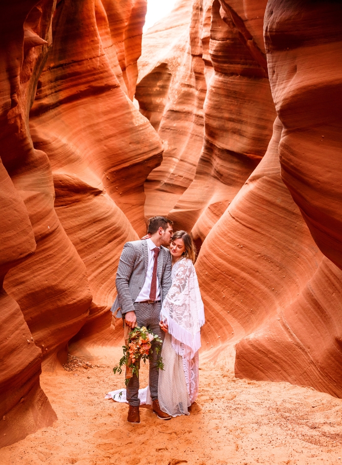 photo of groom kissing bride on the cheek as he holds her bouquet in his hands and she caresses his neck