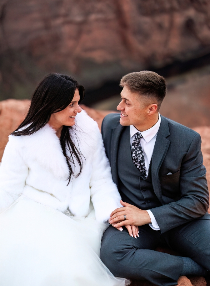 photo of bride and groom holding hands and sitting on a giant red rock