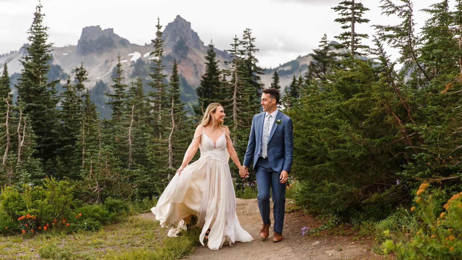 photo of newlyweds holding hands and smiling at each other as they walk down a trail with Mount Rainier behind them