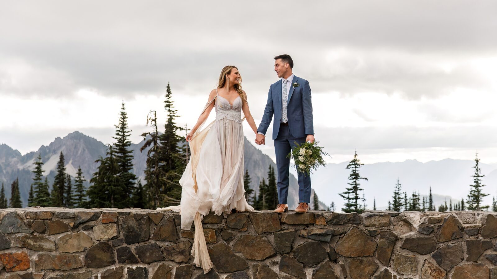 photo of bride and groom standing on stone wall holding hands and looking at each other with Mt Rainier behind them