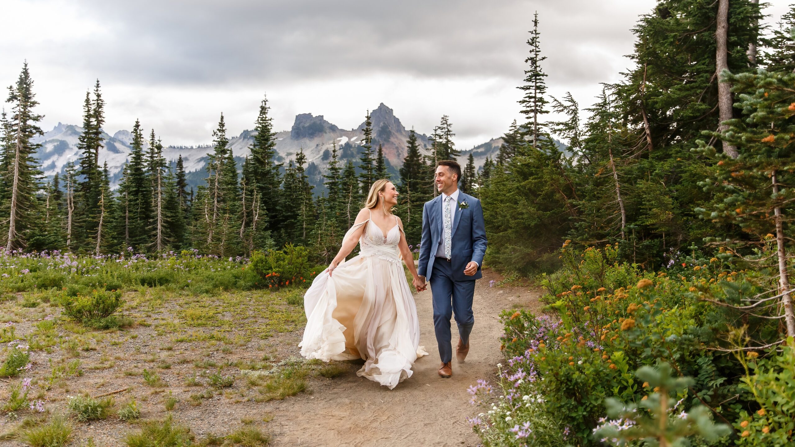 photo of couple holding hands and walking with Mt. Rainier mountain range behind them