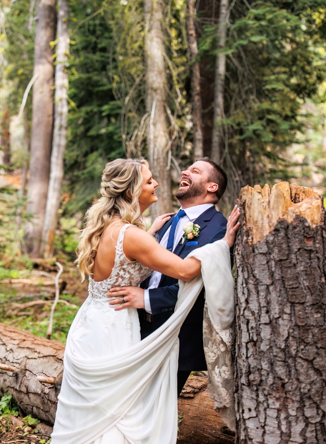 photo of bride and groom laughing while standing next to a tree stump