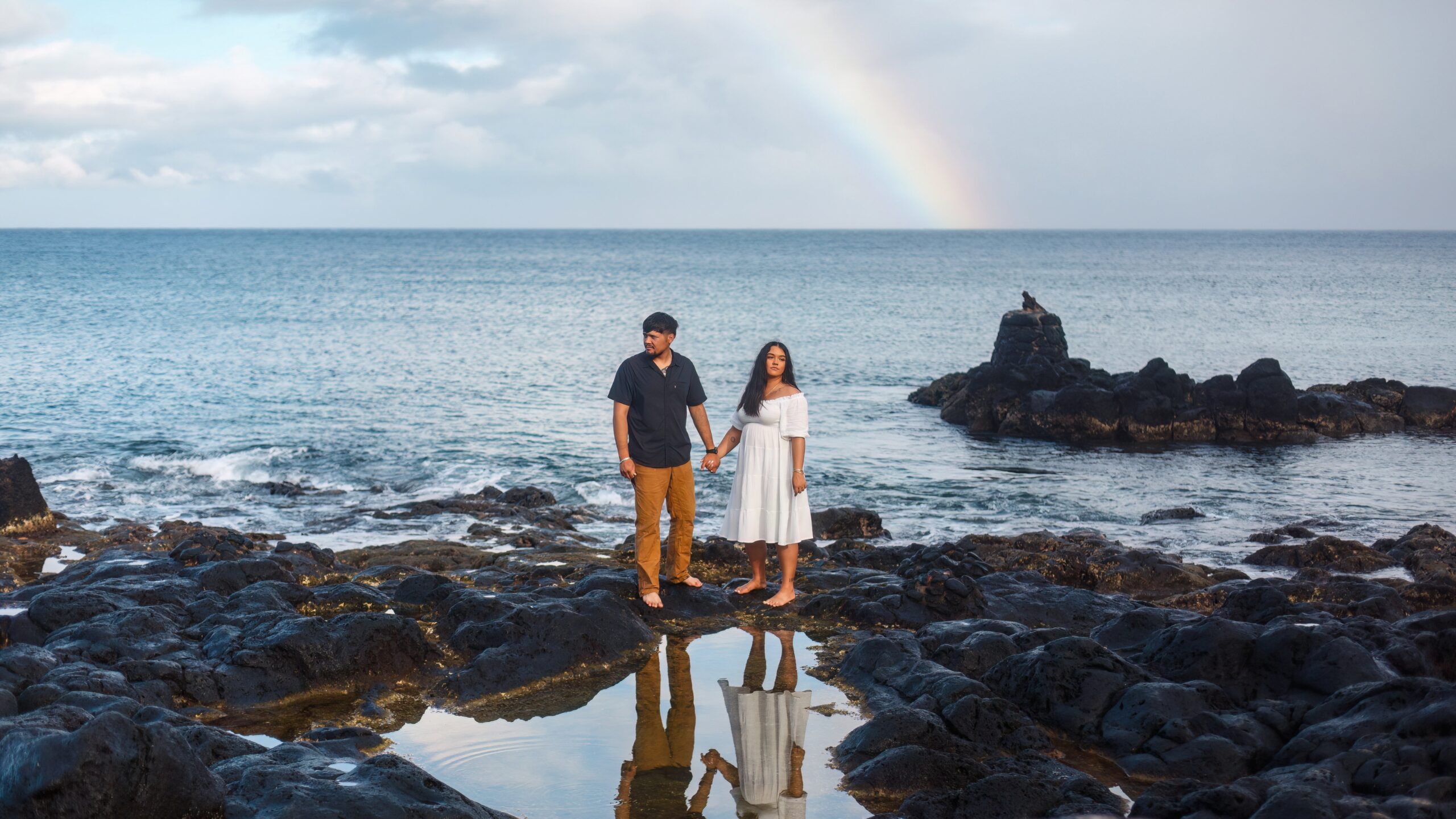 photo of couple holding hands on Hawaii beach with rainbow behind them