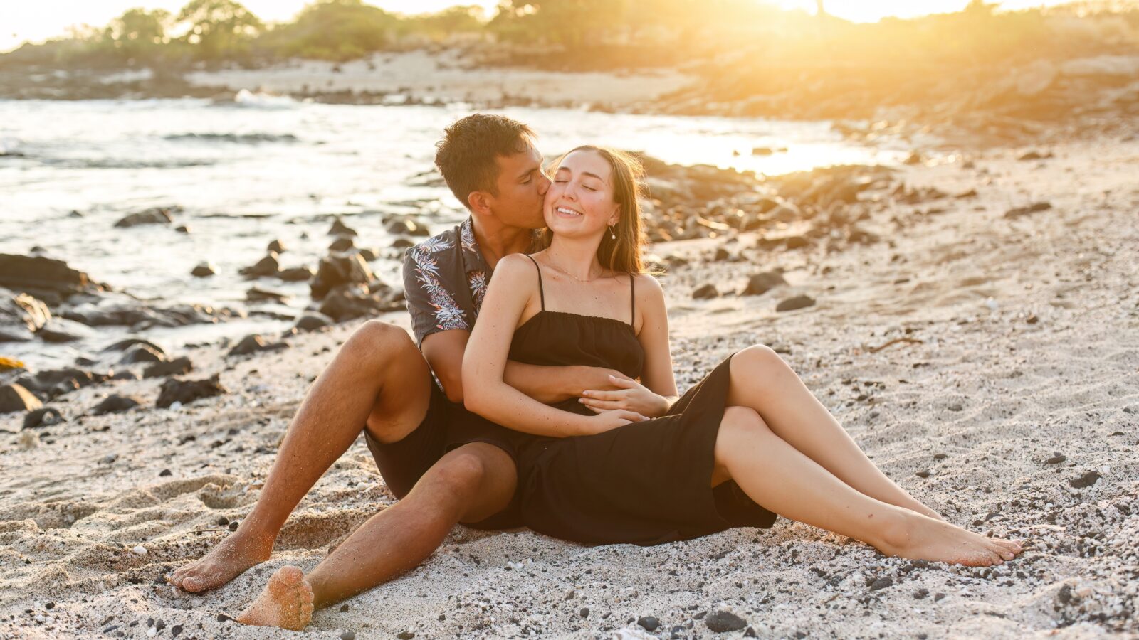 photo of a couple sitting on the beach and a guy holds his girlfriend and kisses her on the cheek as they smile on the beach