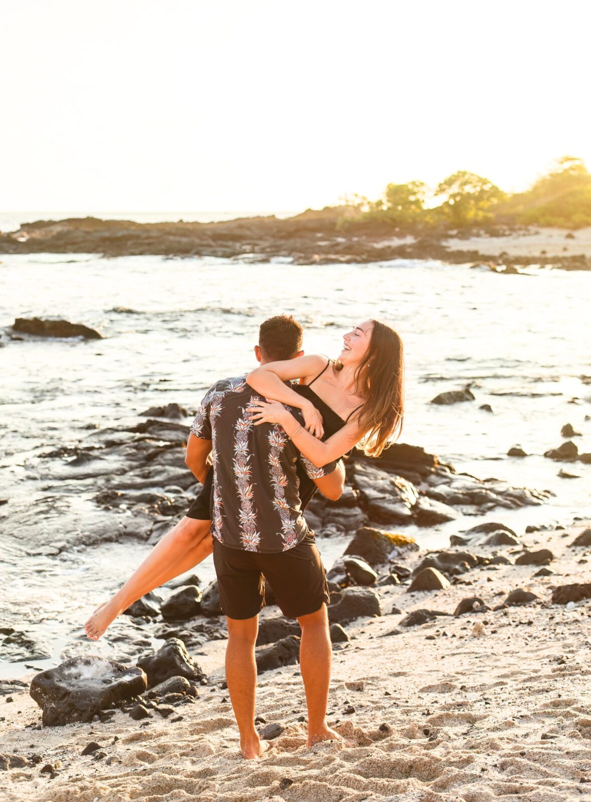 photo of guy carrying his fiancée on the beach as they walk towards to water and she laughs