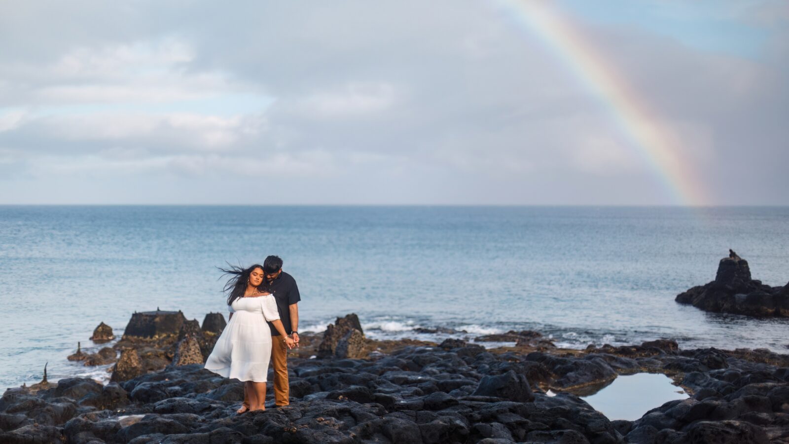 photo of couple holding hand along a lava rock beach with a rainbow behind them