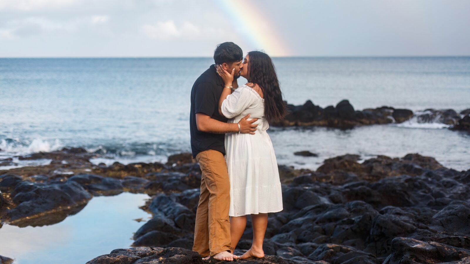 photo of couple standing on lava rock for Hawaii engagement session and kissing with a sunset behind them