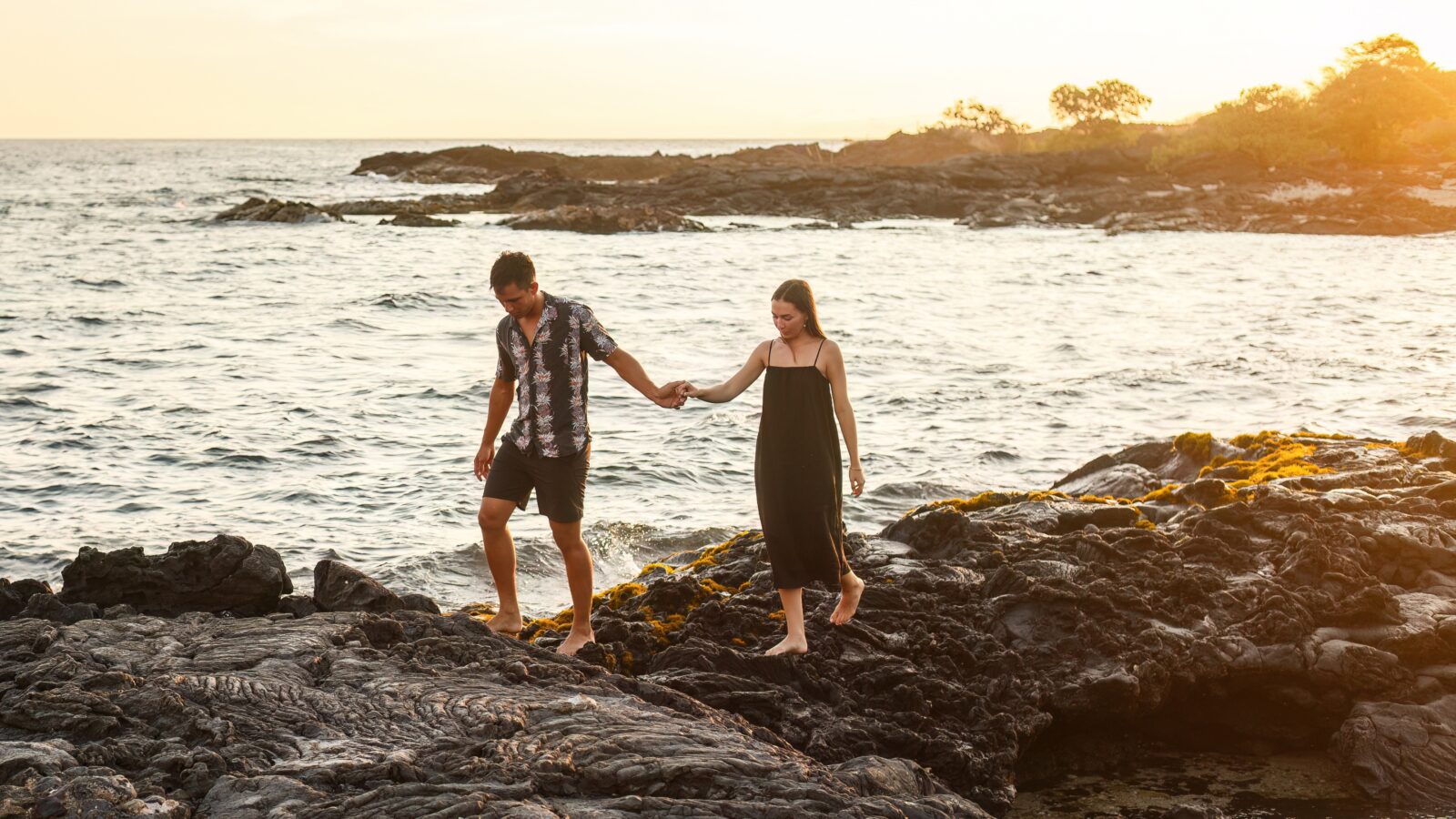 photo of couple holding hands along lava rock beach in Hawaii with sunset behind them