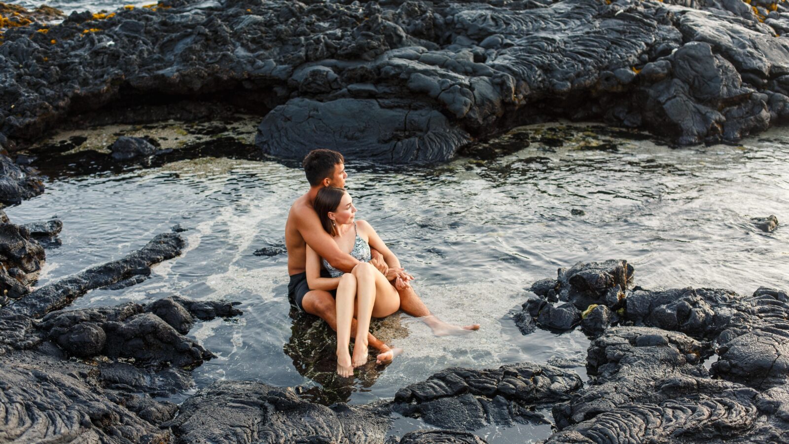 photo of couple sitting in a pool of water surrounded by lava rocks in Hawaii for engagement session