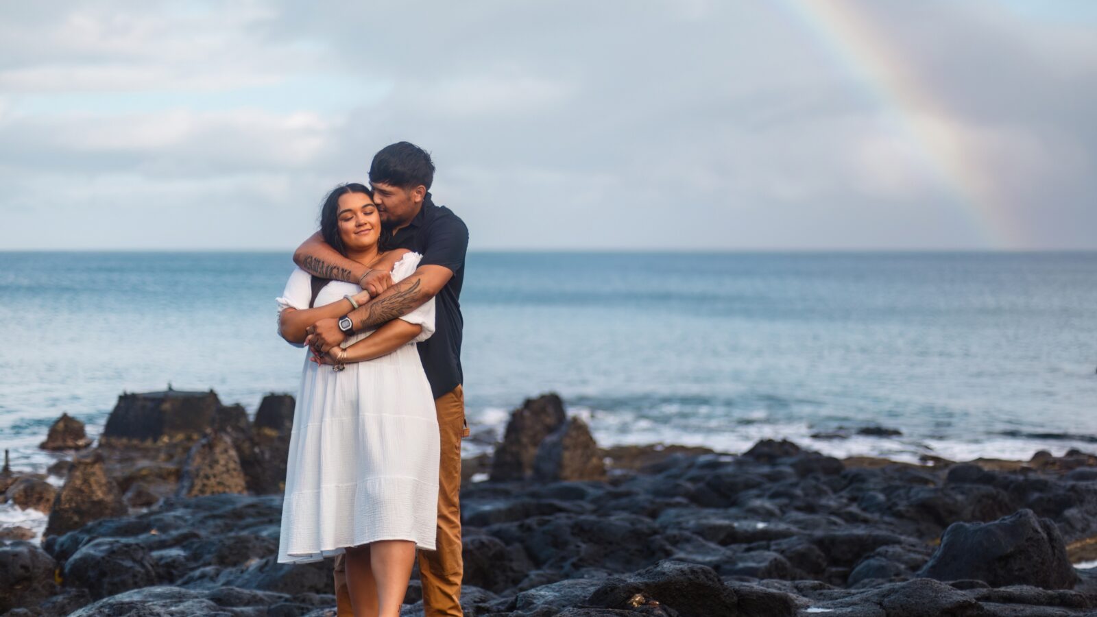 photo of couple embracing on rocky beach in Hawaii with rainbow behind them