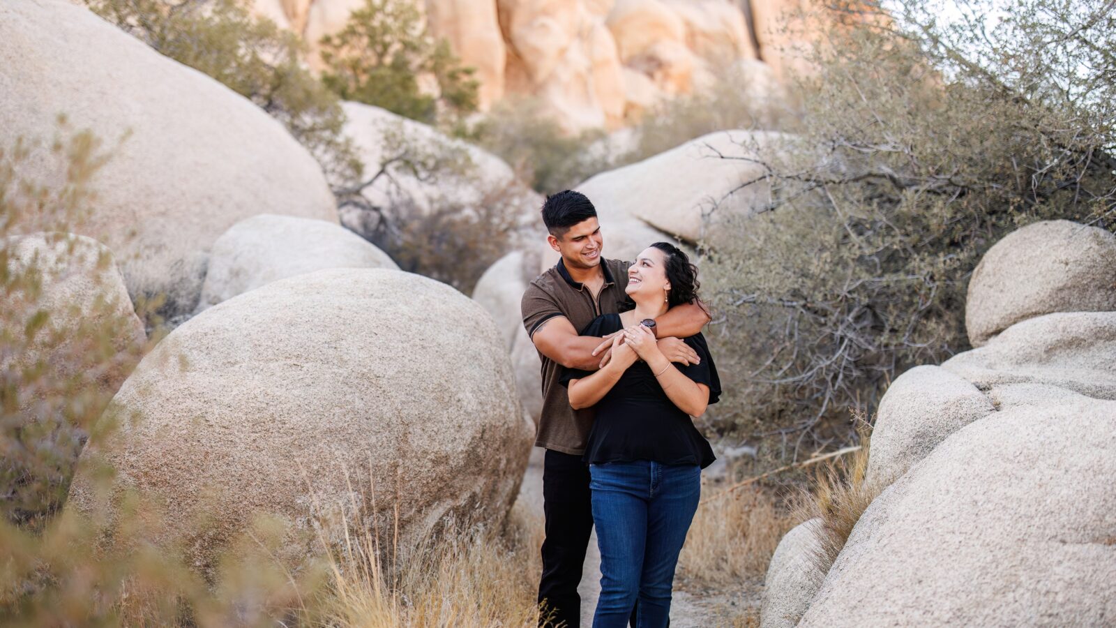 photo of guy standing behind fiancée and smiling at her as she holds his hand