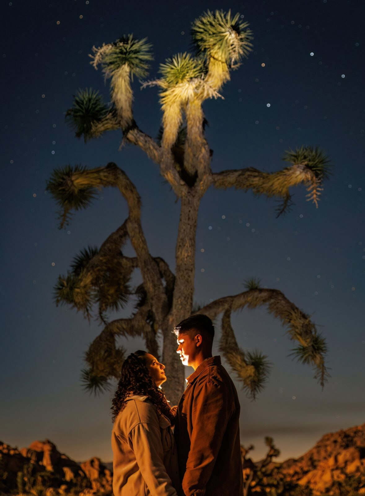 photo of couple looking at each other and smiling in front of a Joshua Tree at night