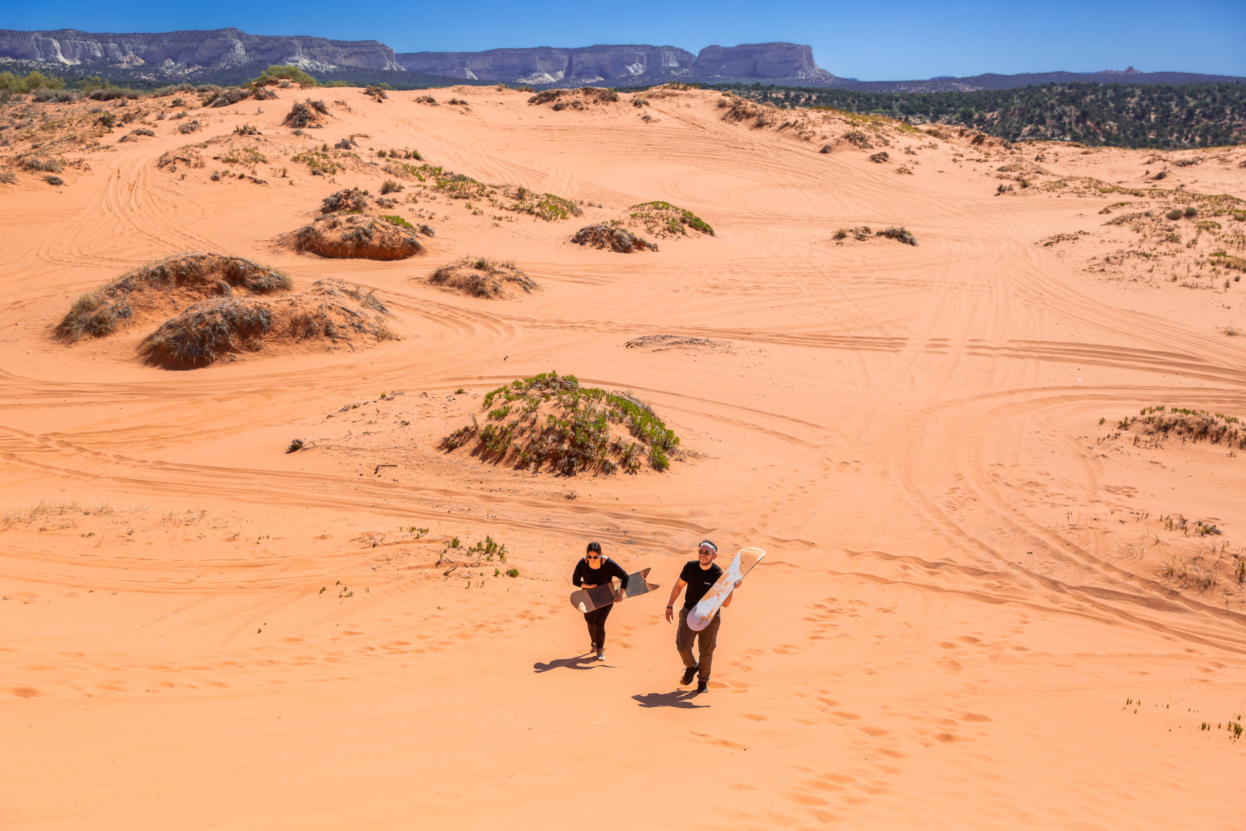 a couple walking up a sand dune with their boards with an incredible view behind them 