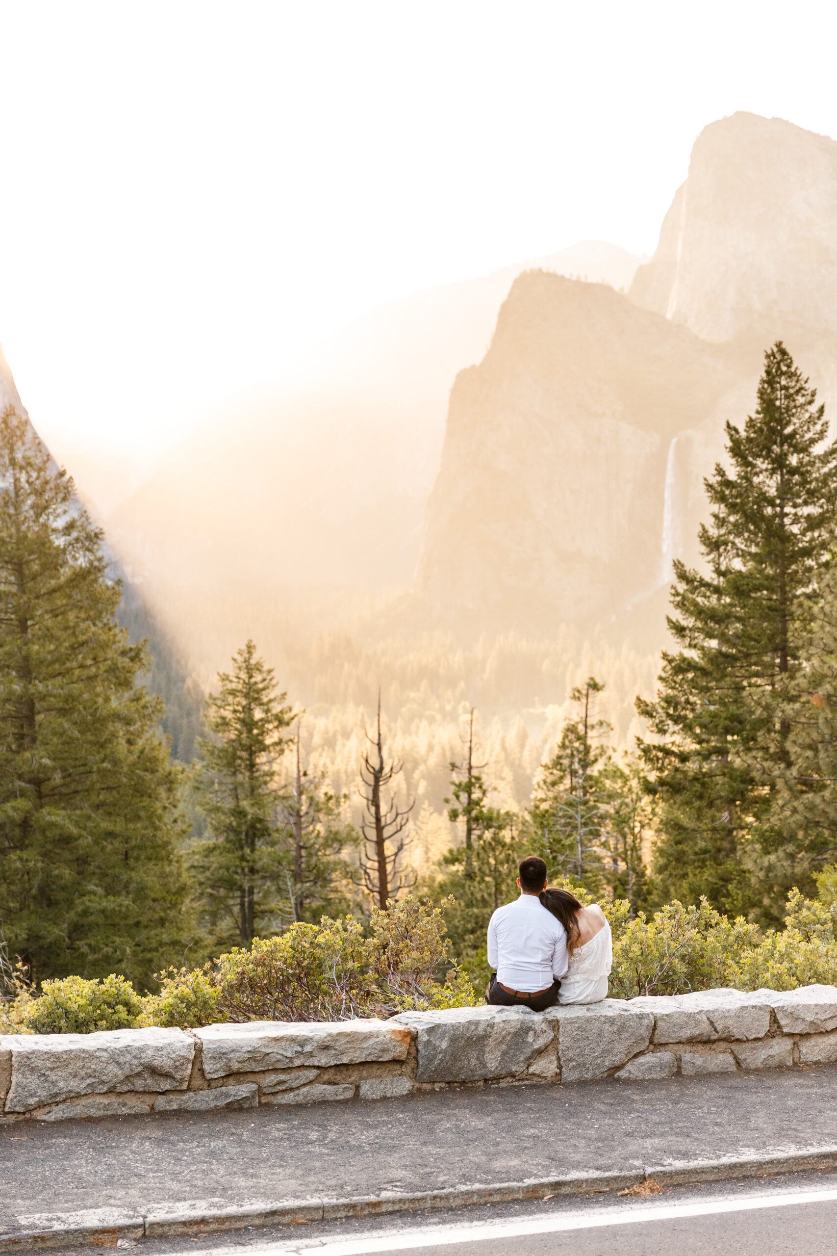 a couple sitting on a ledge during sunrise as the sun shines through the valley 