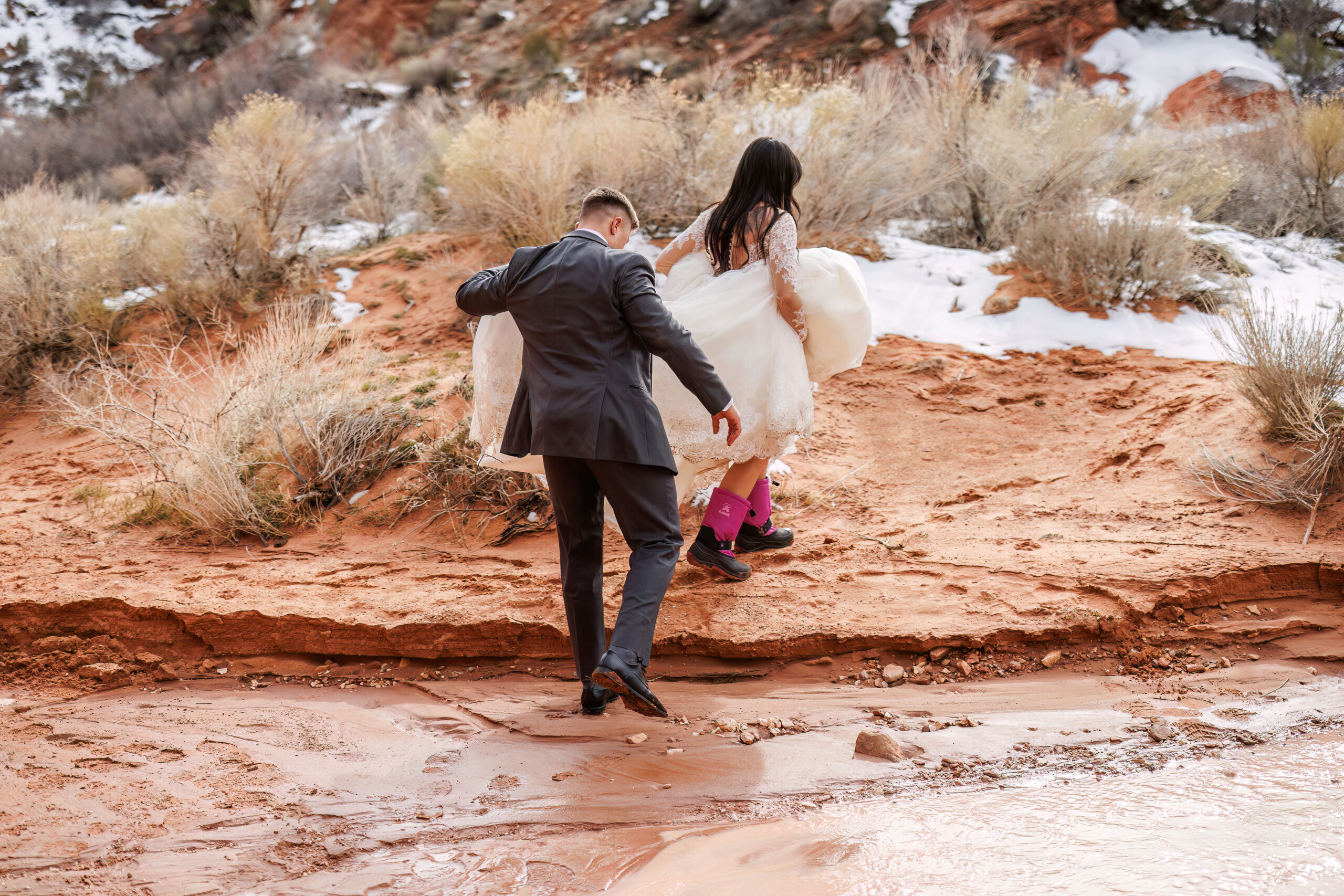 groom holding up bride's dress as they walk through the snowy terrain 