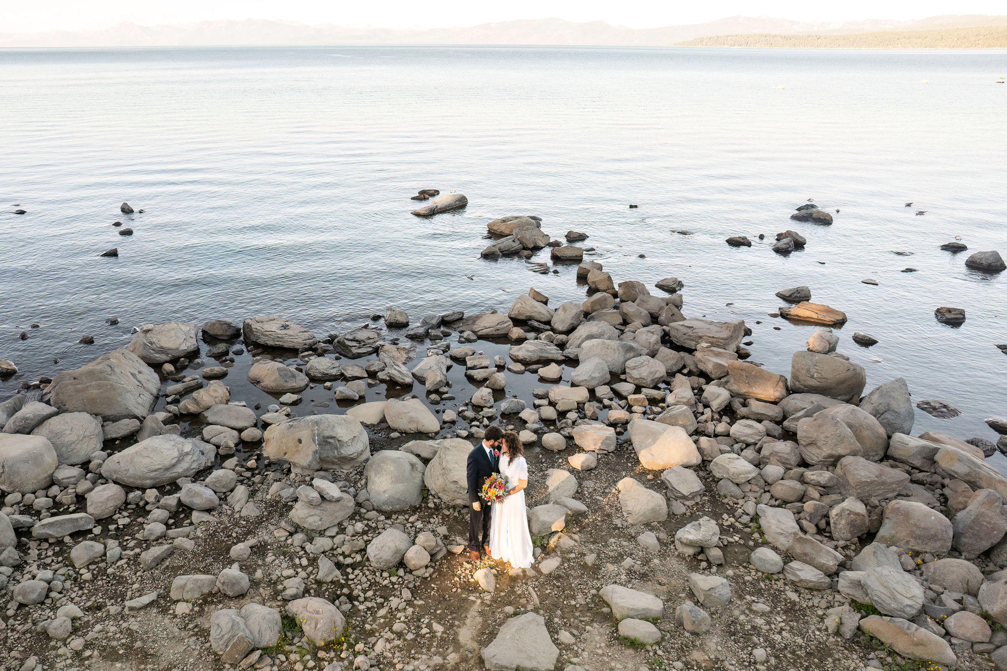 a wide view shot of the bride and groom standing with their foreheads together by the Lake 