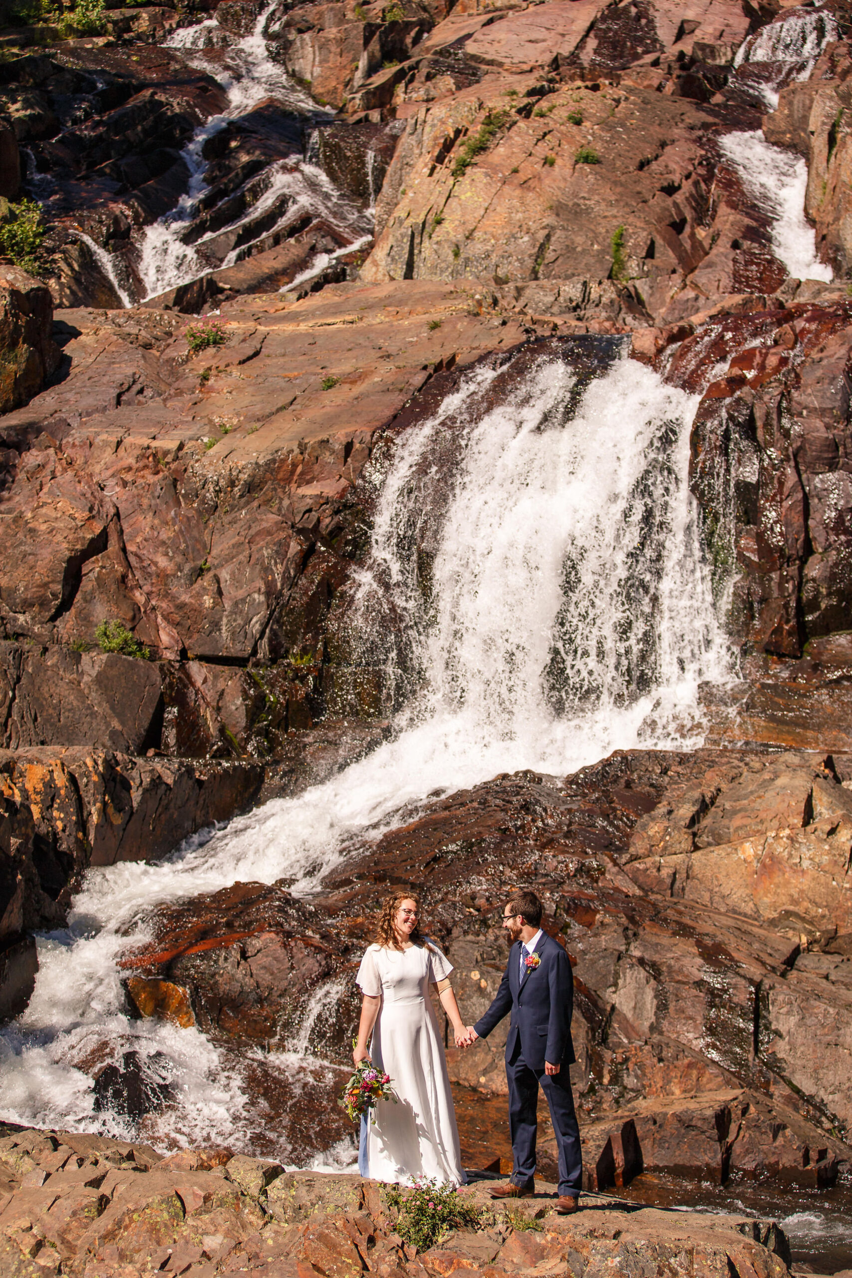 bridal couple holding hands in front of a waterfall during one of their Elopement Activities