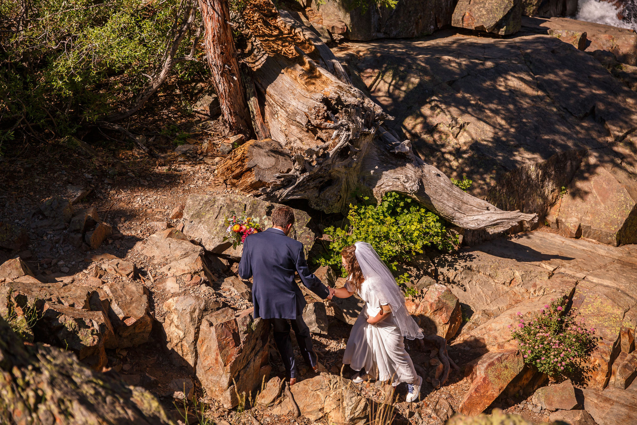 the groom helping the bride walk down through the rocks 