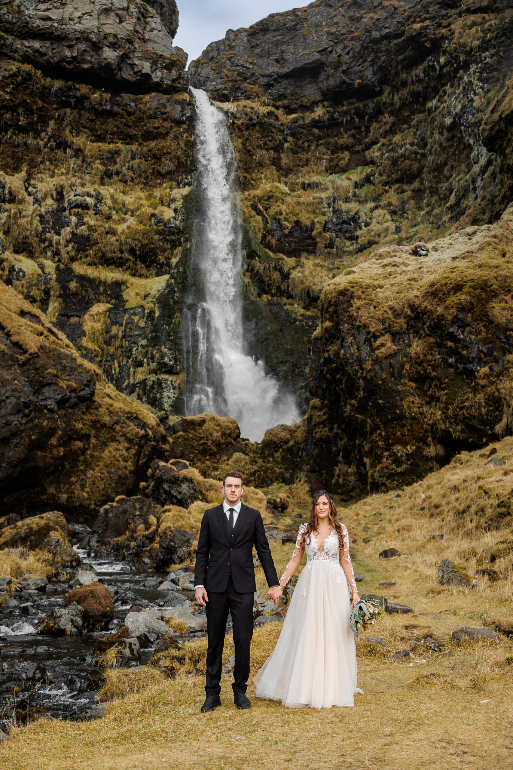 newly weds standing hand and hand in front of the waterfall 