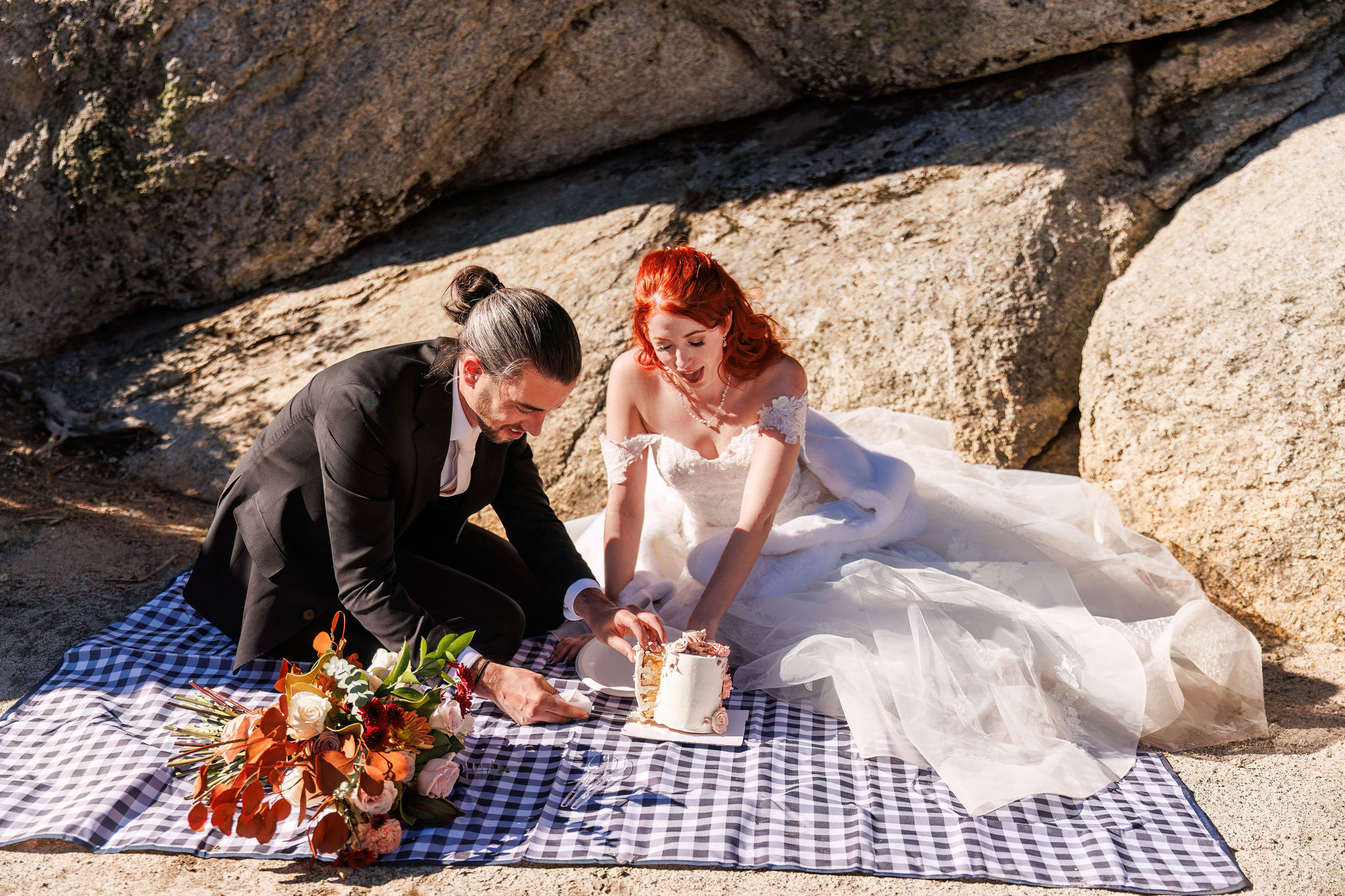 bride and groom sitting on a blanket eating their cake 