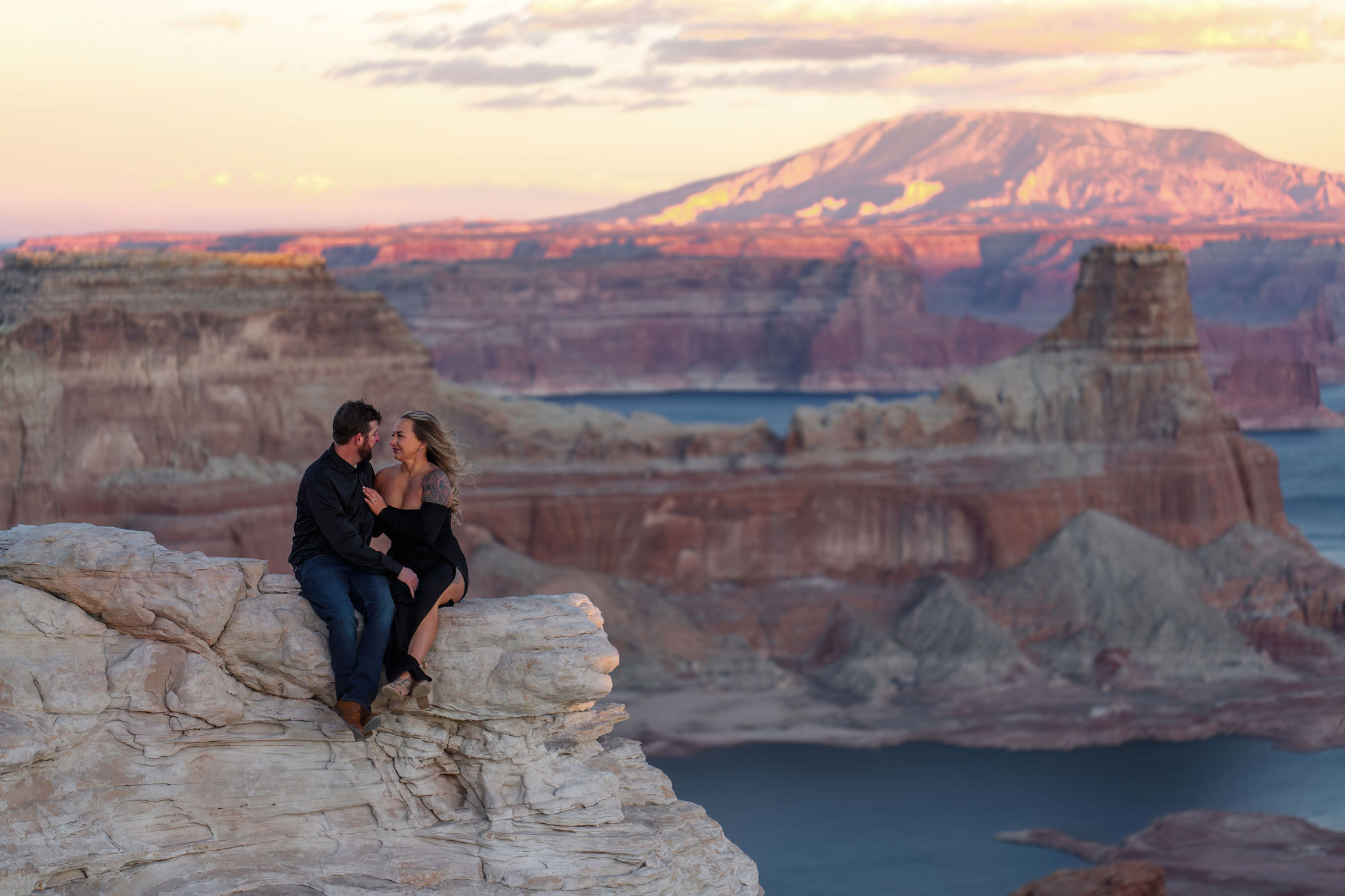 a couple sitting on a rock ledge with a desert lake view in the background 