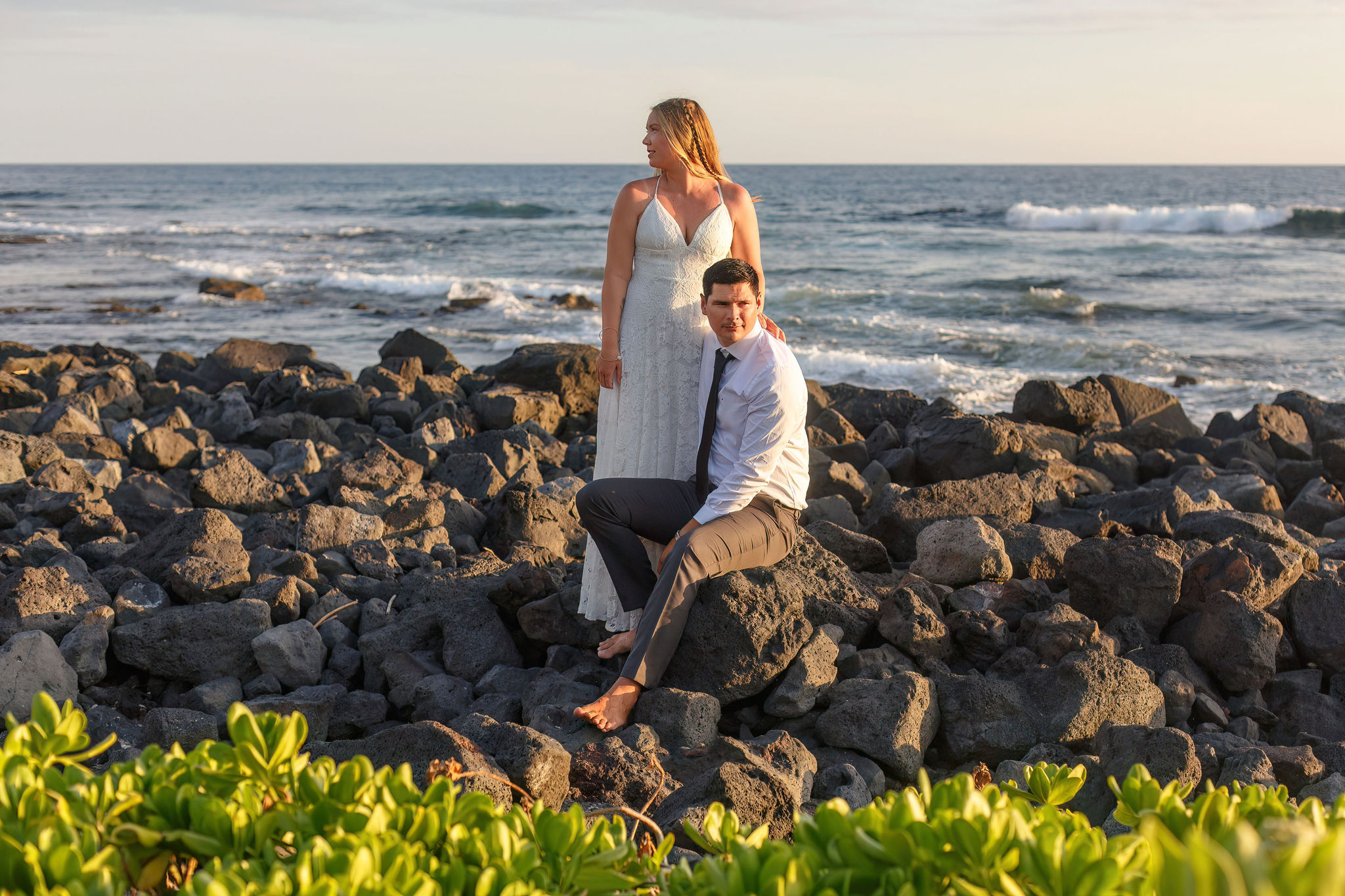 a woman standing next to a man sitting on a rock at the beach 