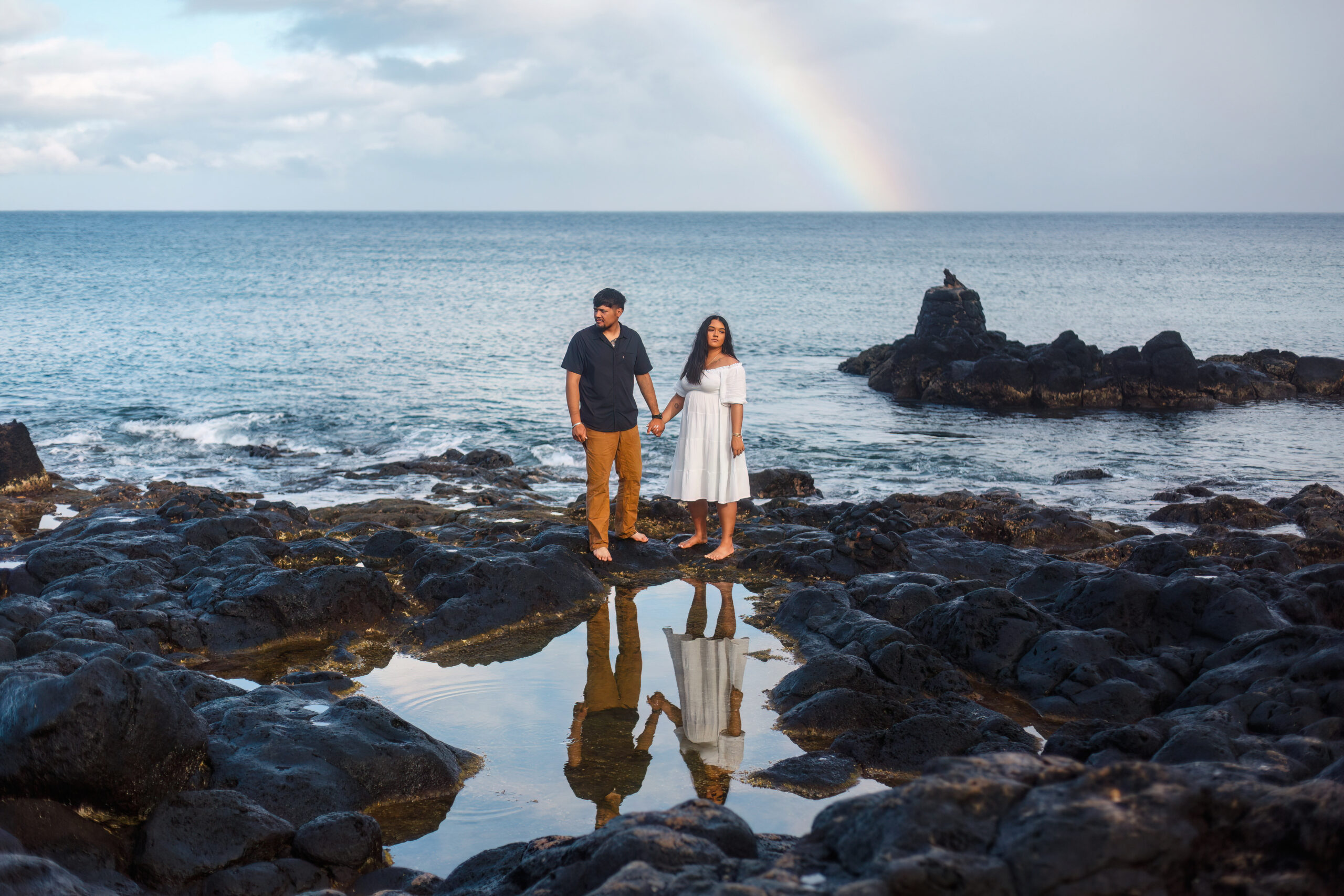 a couple standing on black rocks with a rainbow behind them and their reflection in a pool of water for their Hawaiian elopement