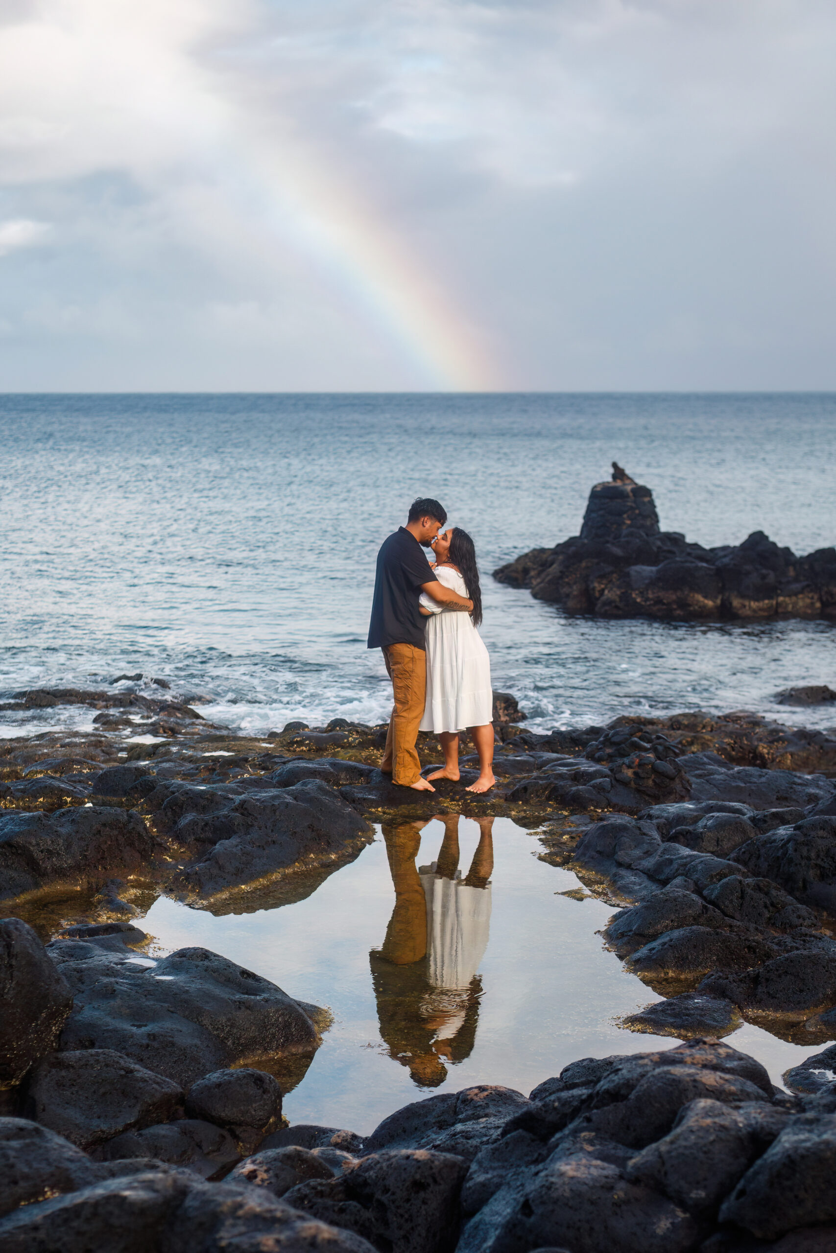 a couple kissing with a rainbow and the ocean behind them