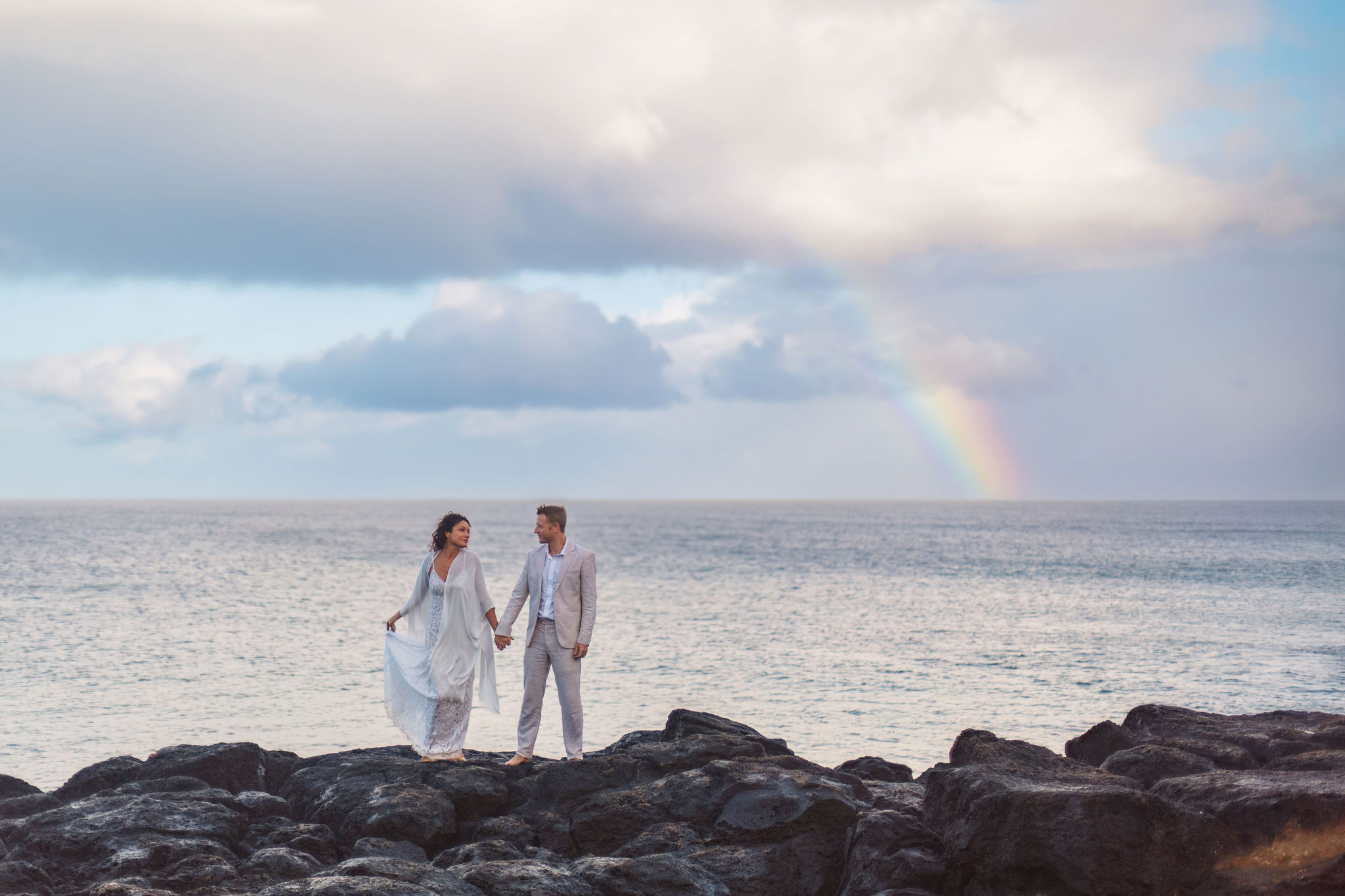 a couple on a rocky beach with a rainbow behind them during a Hawaiian elopement