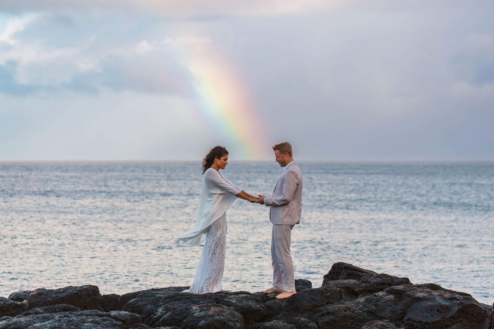 a couple standing on black rocks, holding hands, with a rainbow in the background