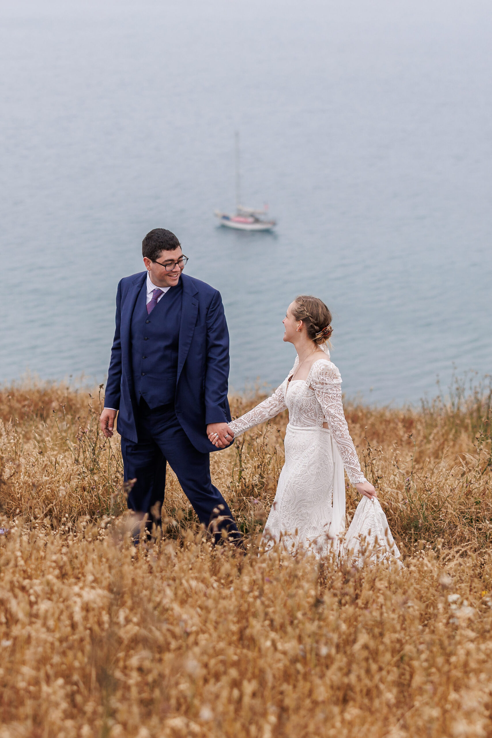 newly weds walking through the tall grass together with the ocean in the background