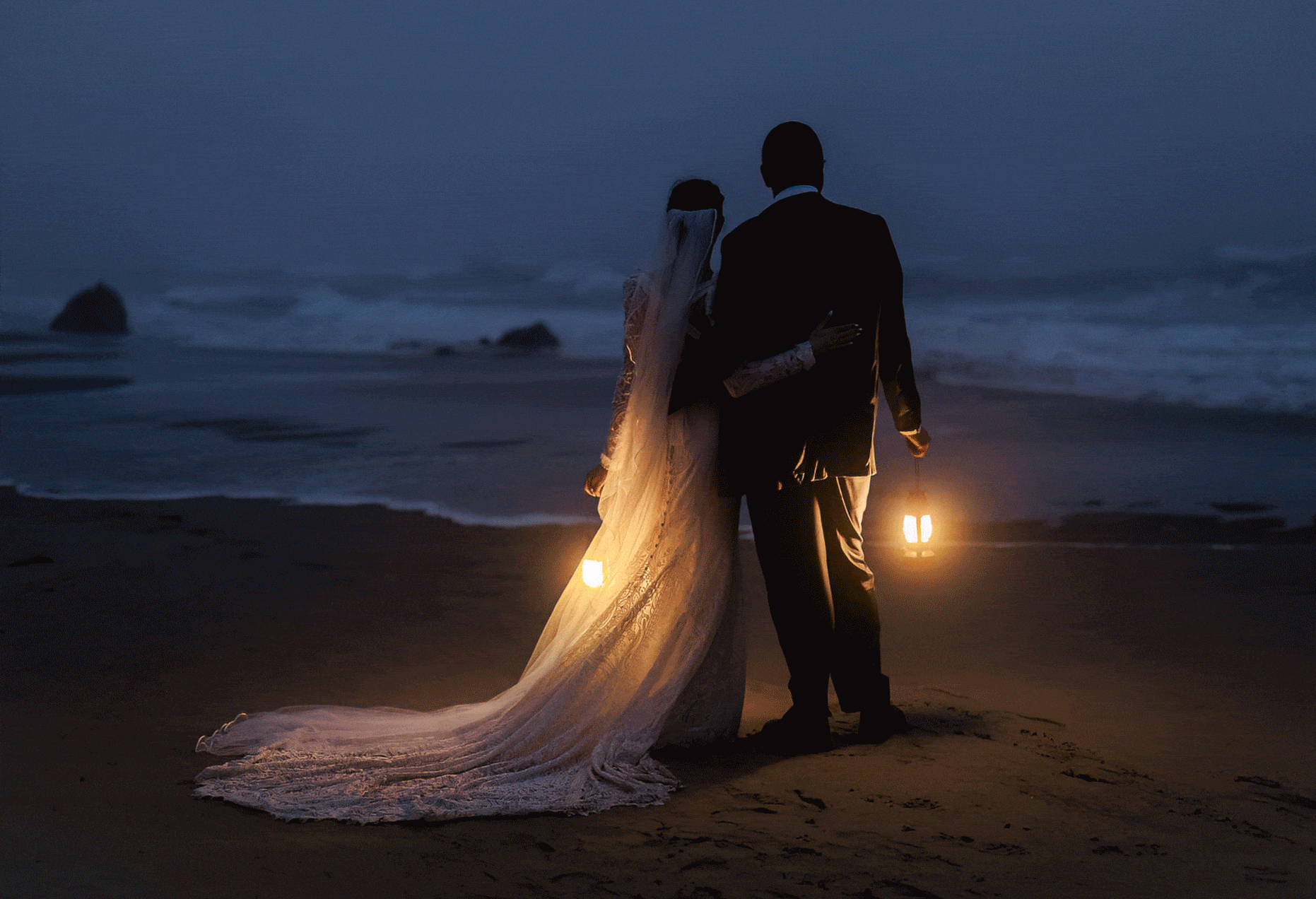 newly weds standing on the beach at dark, holding lanterns, looking out into the ocean
